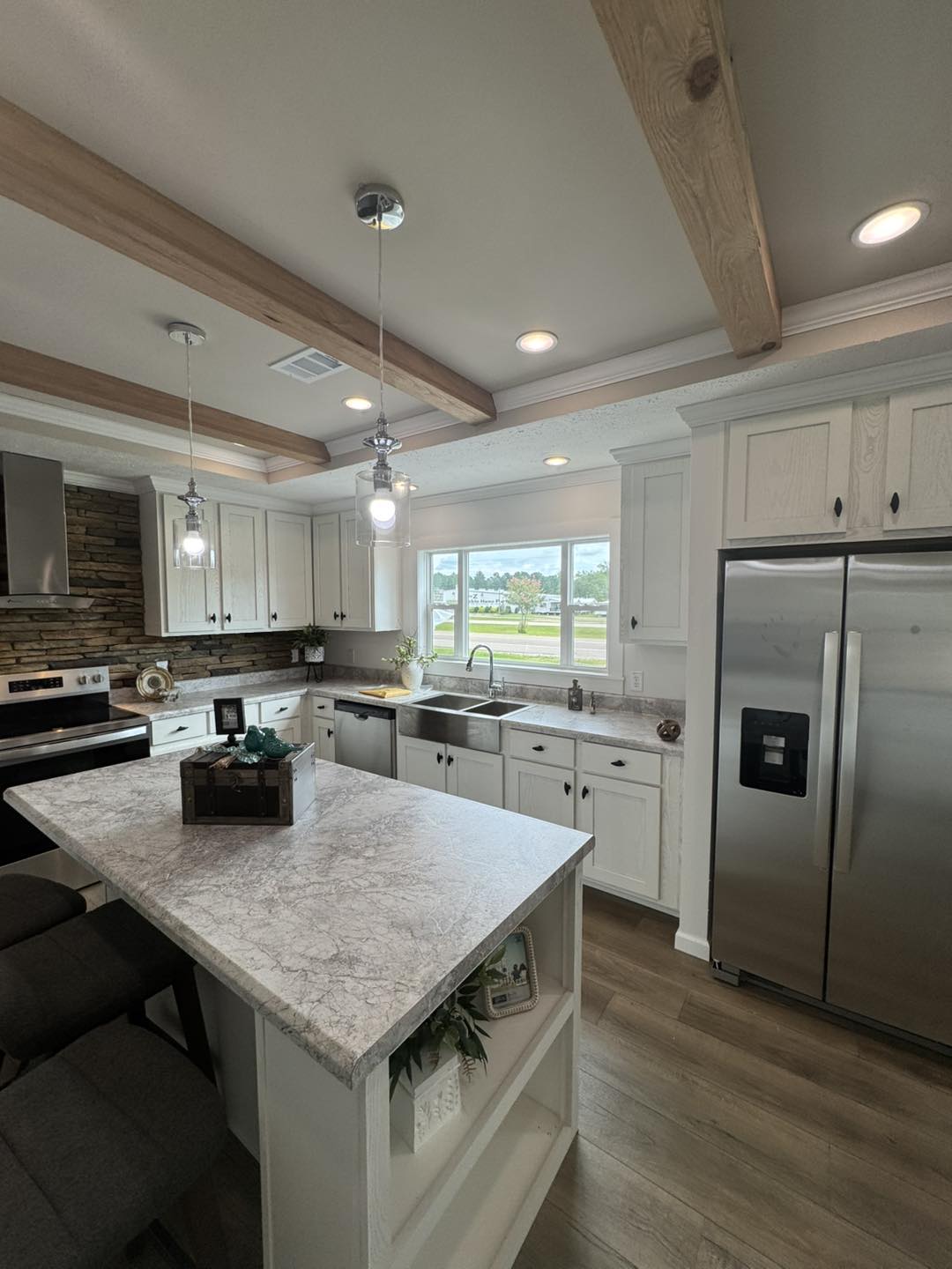 Modern kitchen with white cabinets, stainless steel appliances, and a large window above a farmhouse sink. Features a stone backsplash, island with bar stools, and wood beam ceiling for a cozy, rustic feel. Natural light brightens the space.