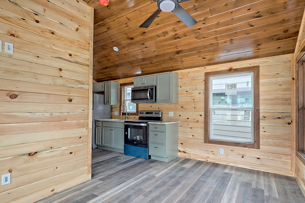 Cozy kitchen in a cabin with pine walls and ceiling, modern gray cabinets, stainless steel appliances, and a ceiling fan. Bright, rustic, inviting.