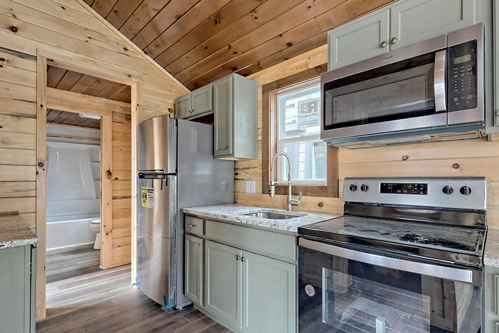 Cozy kitchen with wooden walls and ceiling, featuring sage green cabinets, stainless steel appliances including a fridge, stove, and microwave, with a glimpse of a bathroom.