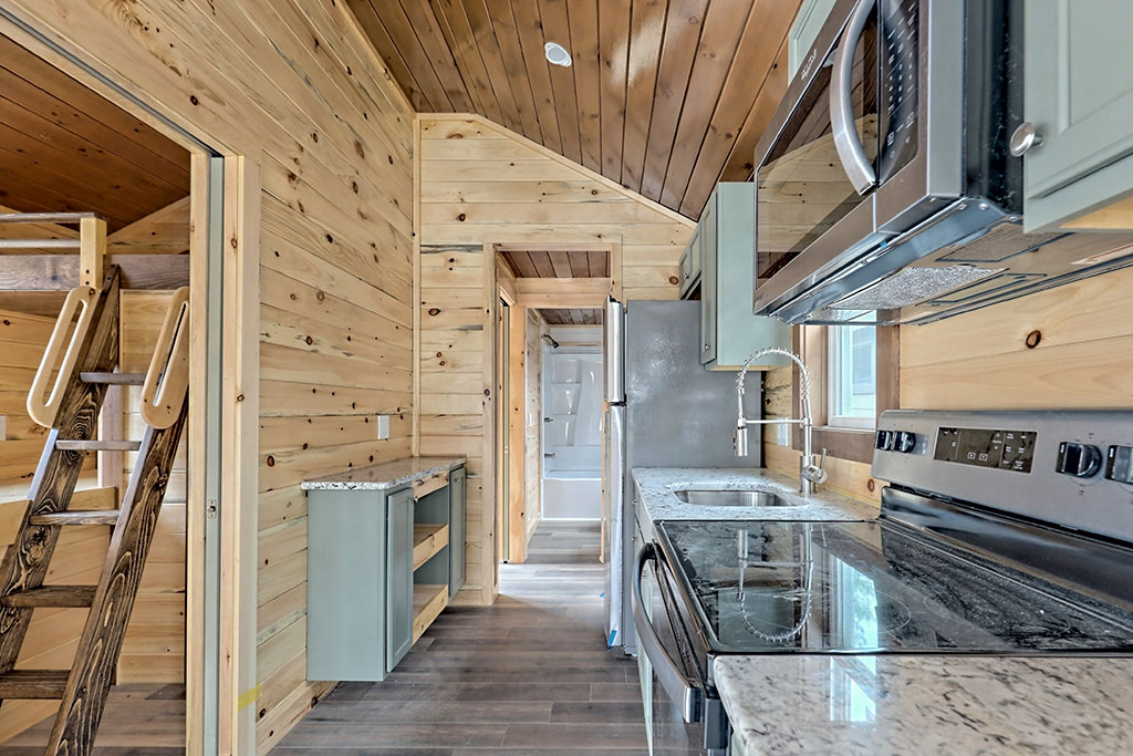 Compact kitchen with stainless steel appliances in a wood-paneled tiny home. Ladder leads to loft, creating a cozy, rustic atmosphere.