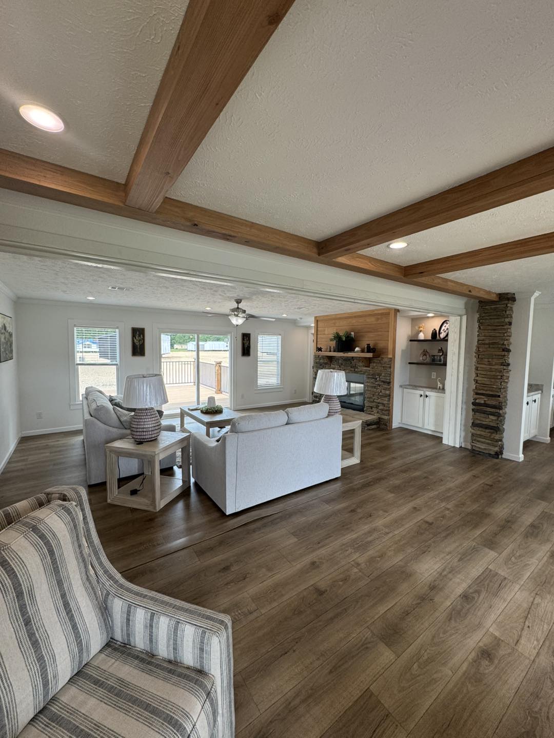Spacious living room with wooden beams and flooring, featuring a cozy sitting area with white sofas and a stone fireplace. Large windows offer ample natural light.