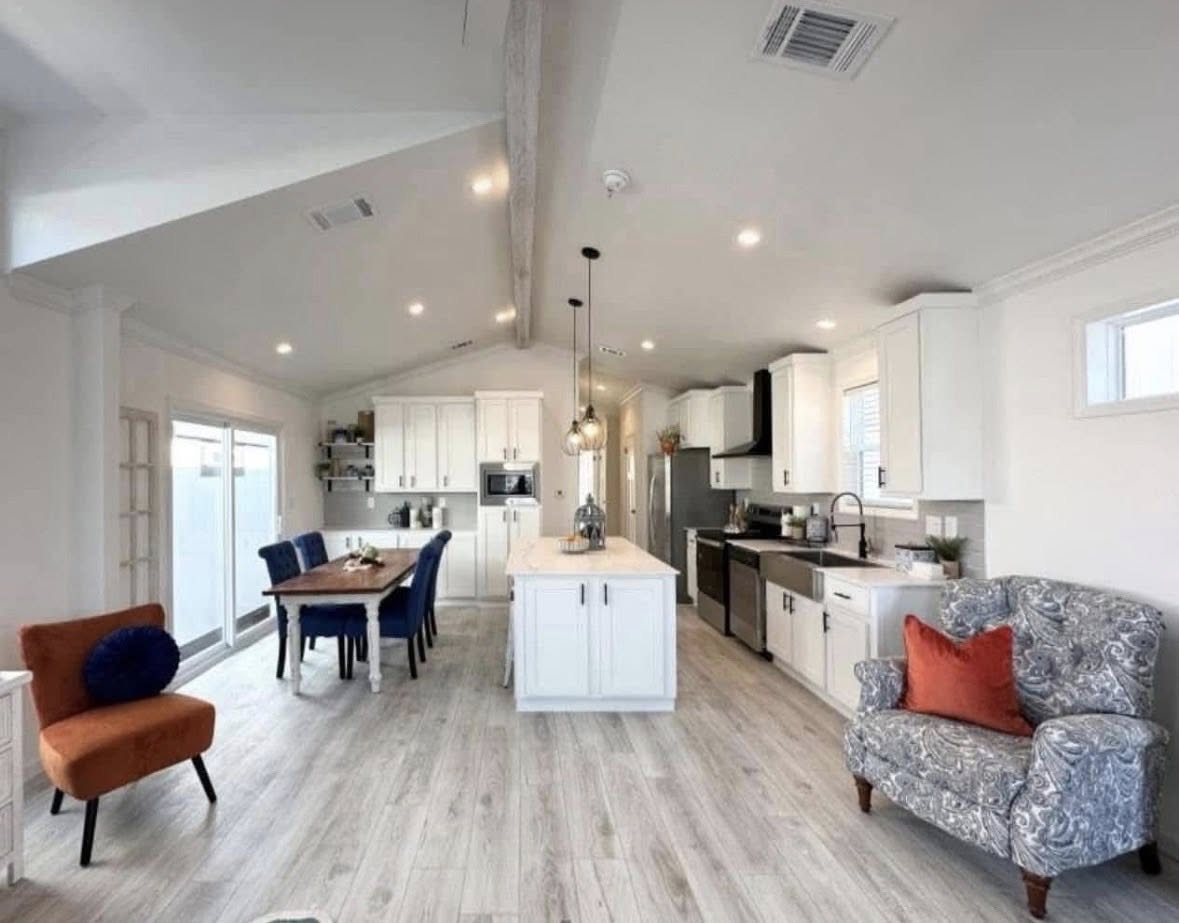 Spacious modern kitchen and dining area with white cabinetry, wood flooring, and pendant lighting. Features a blue-gray armchair, orange accent chair, and blue dining chairs. Cozy and stylish ambiance.