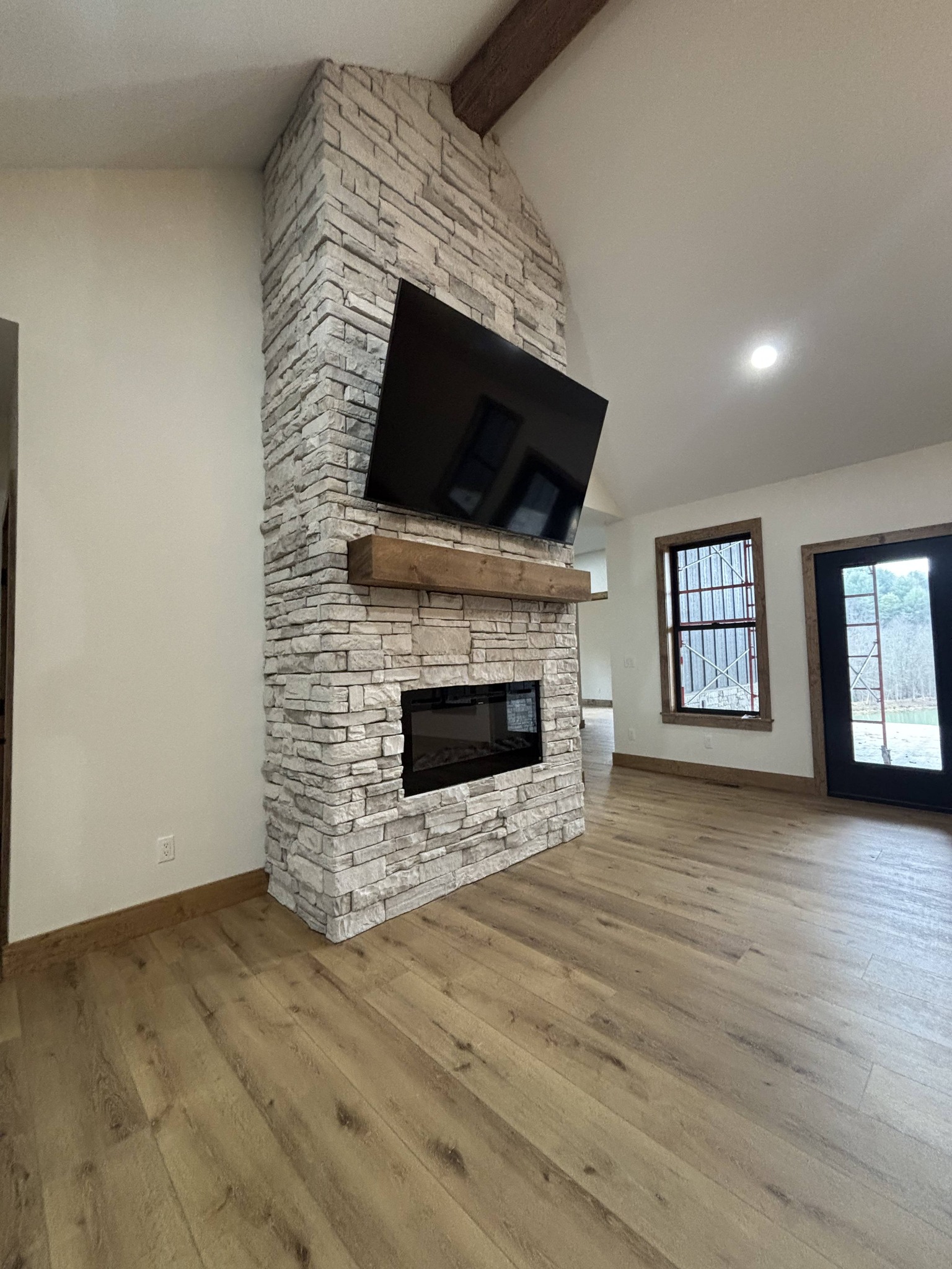 Living room with a tall, white stone fireplace. A large TV is mounted above a wooden mantel. The space features wooden floors and a large window, creating a cozy atmosphere.