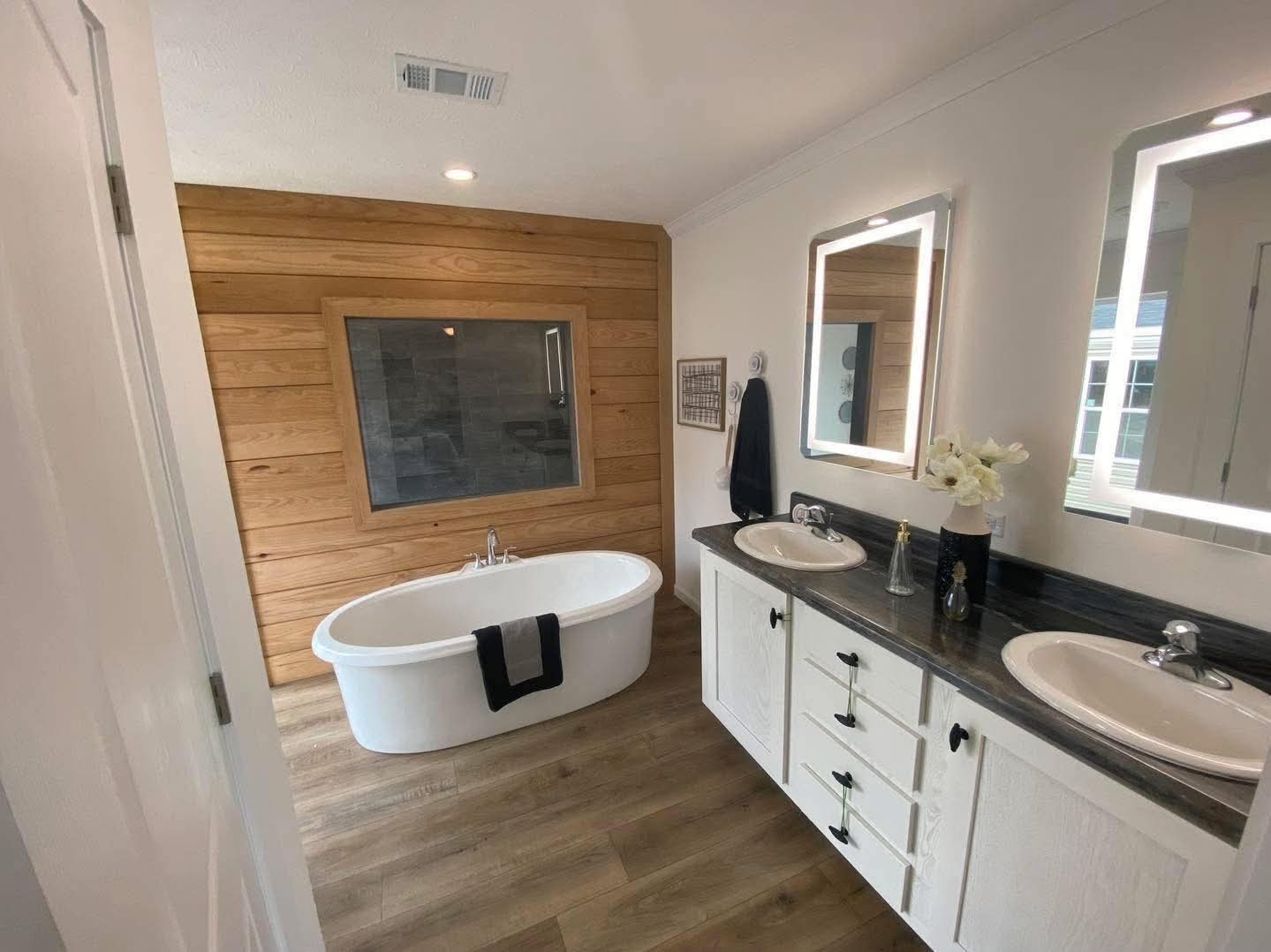 Modern bathroom with a freestanding white tub, wood accent wall, double sink vanity, illuminated mirrors, and a towel on the tub. Warm, inviting ambiance.