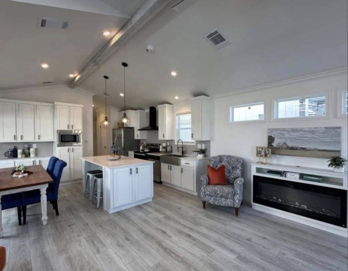 Modern open-concept kitchen and living area with white cabinetry, wood flooring, a large island, pendant lights, and an armchair with an orange pillow.