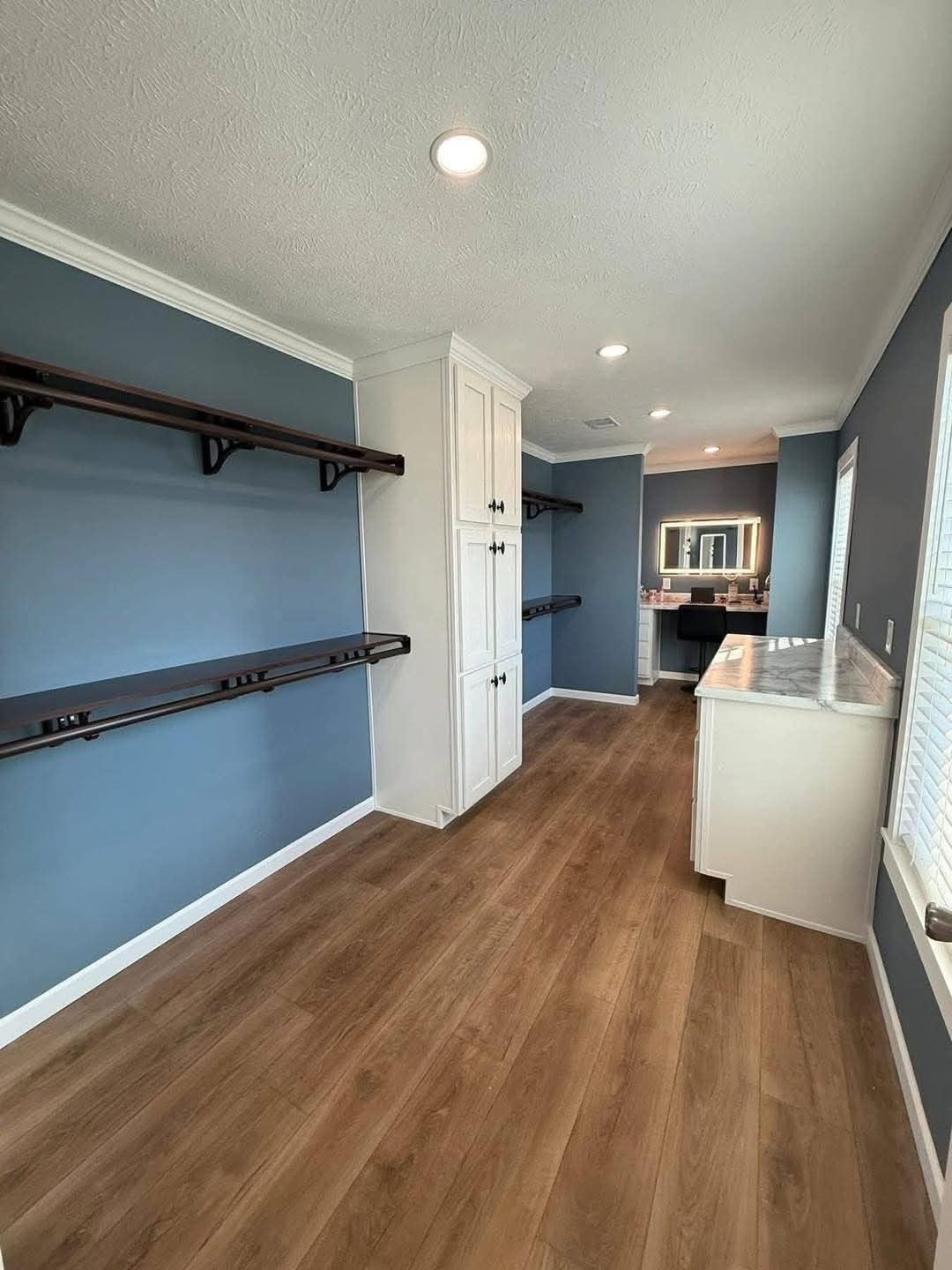 A spacious, modern closet with blue walls, wooden flooring, open shelves, and a white cabinet. Bright lighting and a vanity area in the distance.