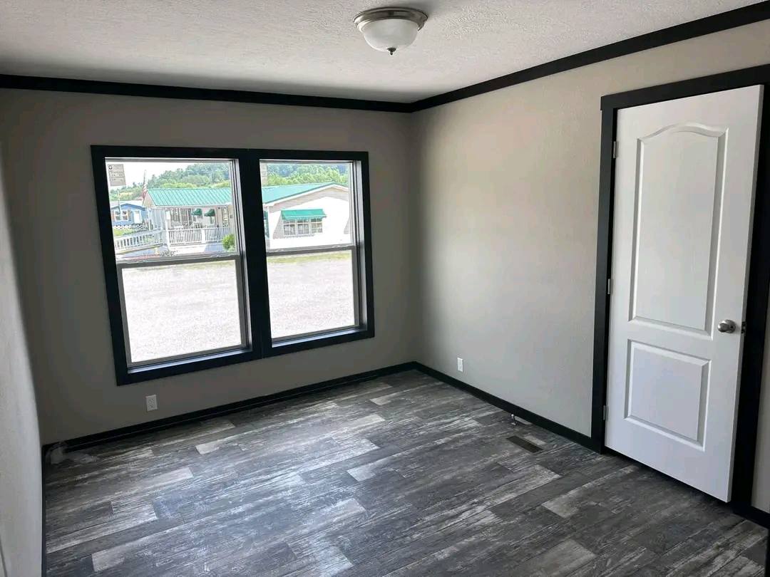 Empty room with gray walls, dark trim, and wood-like flooring. Two large windows let in natural light, revealing an outside view of houses. Simple, neutral tone.