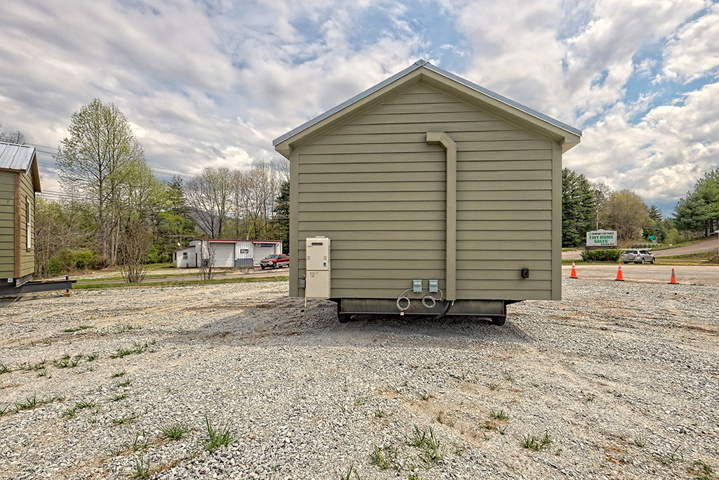 A small, green wooden structure with a gabled roof on a gravel lot. Trees and a cloudy sky in the background convey a rural, peaceful setting.