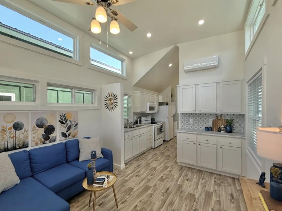 A modern living space with a blue sectional sofa, abstract floral wall art, and a wooden coffee table. White kitchen cabinets and hardwood floors create a cozy atmosphere.