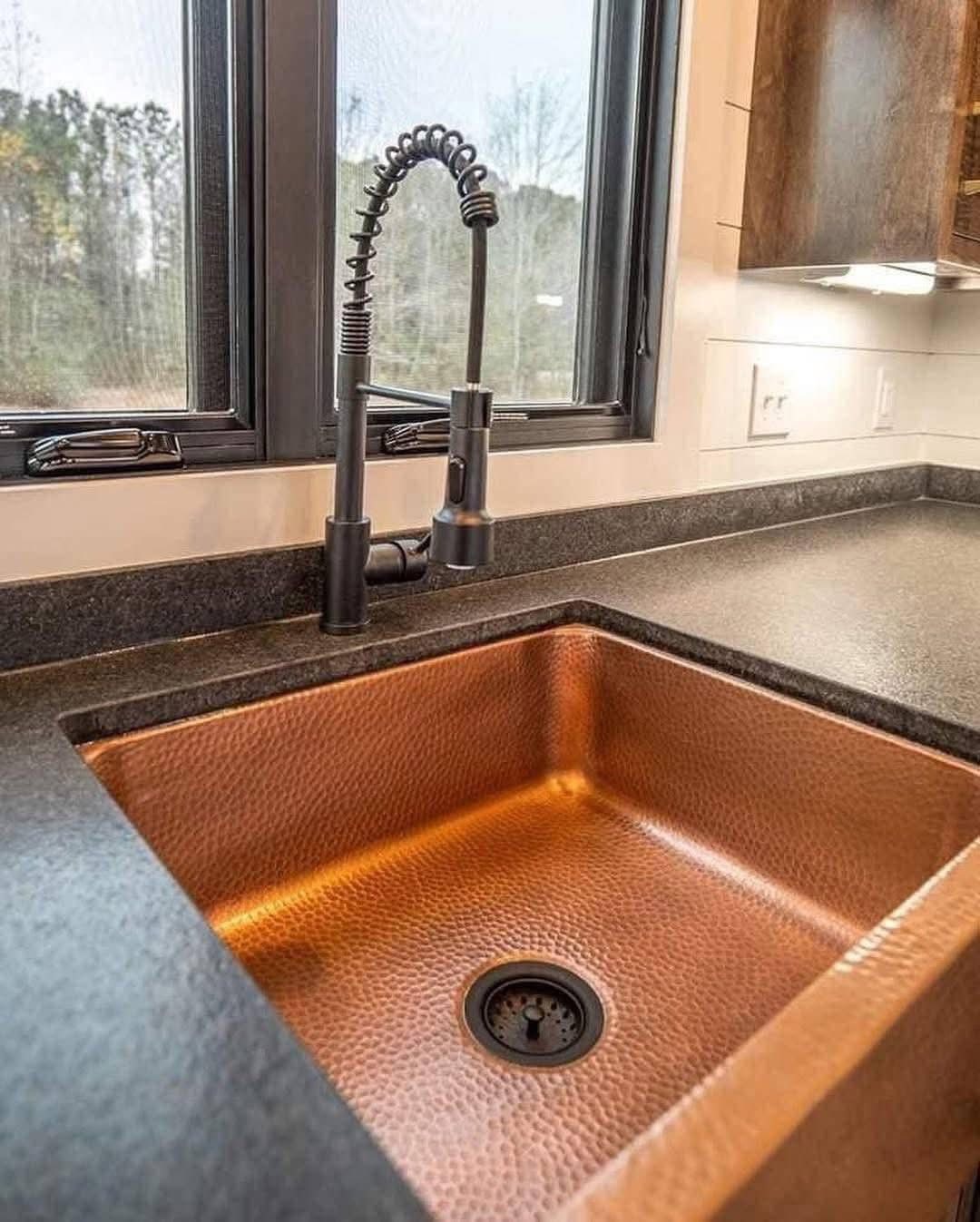 Copper kitchen sink with a textured finish, complemented by a matte black, spring-neck faucet. Set against a dark countertop and large windows.