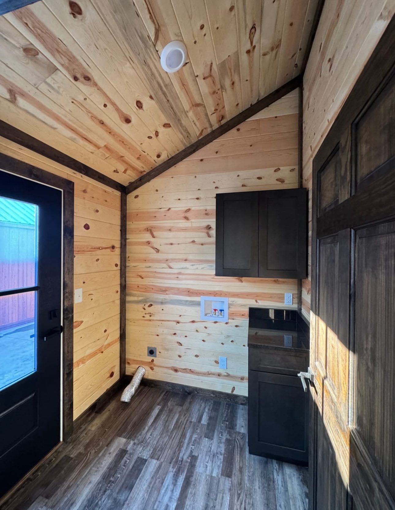 Small wooden room with pine walls and ceiling, featuring a dark wooden door on the left and cabinets on the right. A cozy, rustic interior.