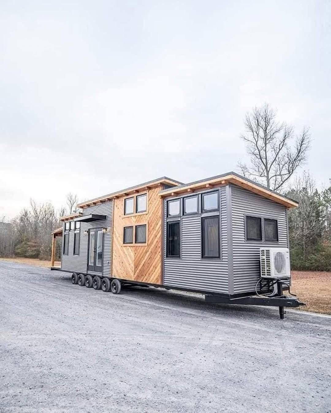 A modern tiny house on wheels featuring geometric wood and metal paneling sits on a gravel road. The surrounding area is open with bare trees, suggesting a winter setting.