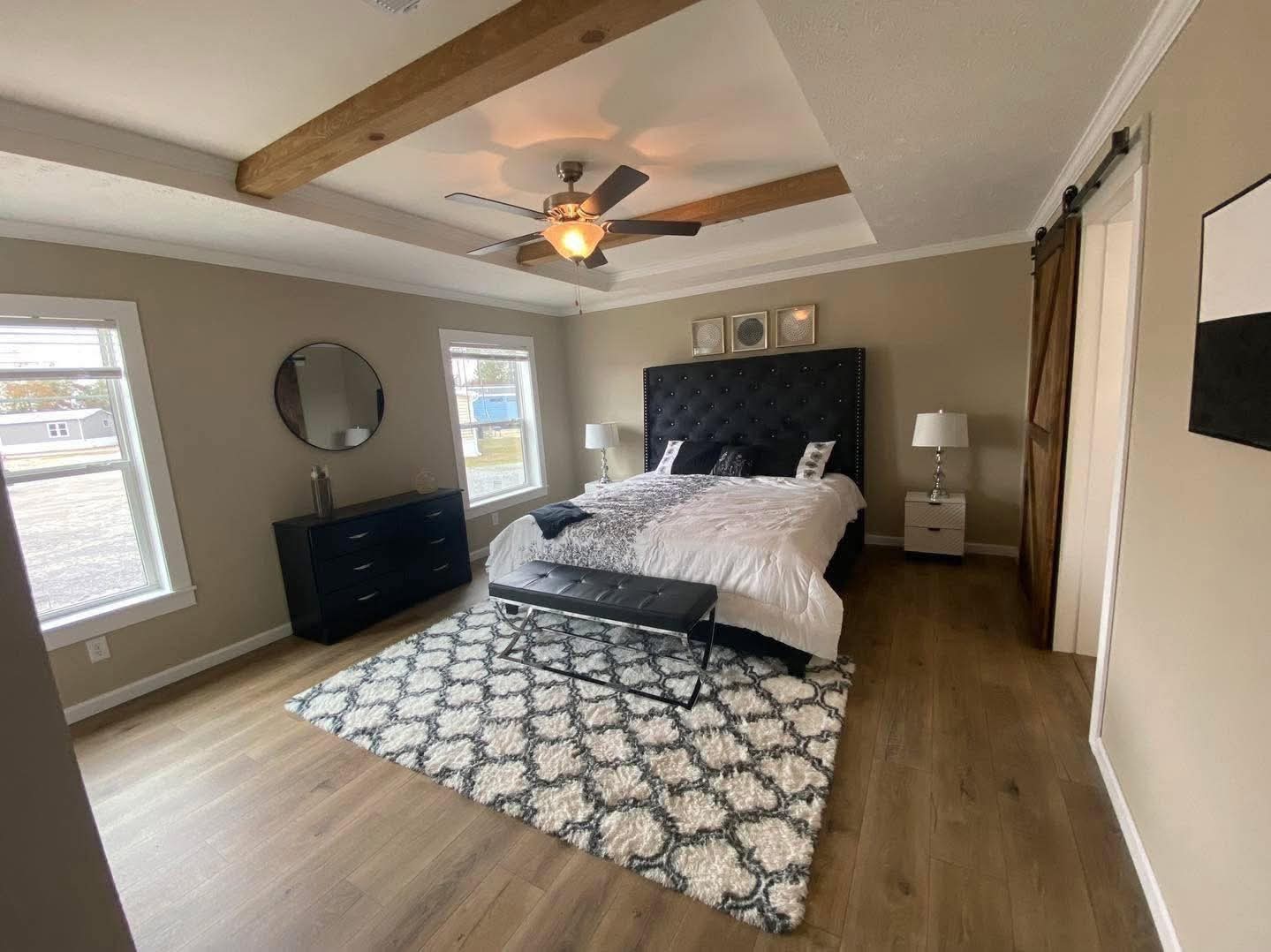 Spacious bedroom with wood flooring, a dark tufted headboard, and a soft, patterned rug. Features a ceiling fan, dresser with round mirror, and barn door.