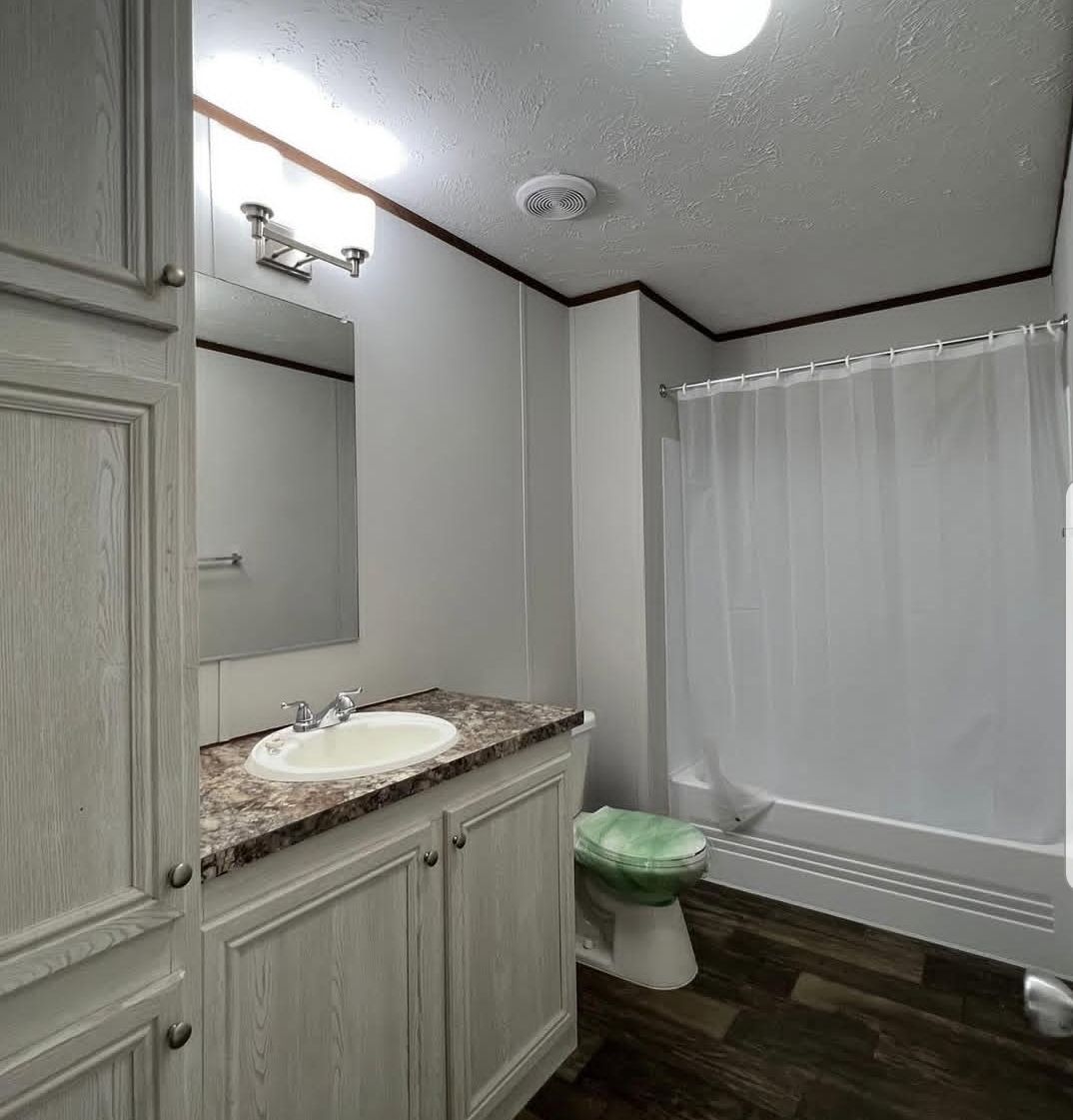 Small bathroom with wood flooring, white cabinets, marble countertop, and sink. Shower with white curtain and toilet. Soft lighting creates a clean, cozy feel.