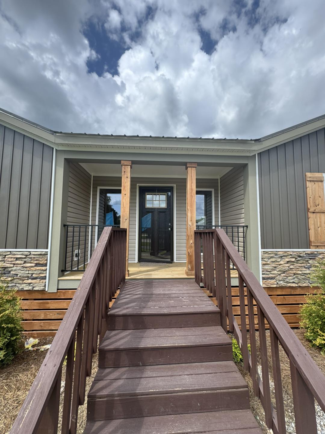 A brown wooden staircase leads to a modern front door between stone-trimmed pillars on a cloudy day, creating a welcoming and serene entrance.