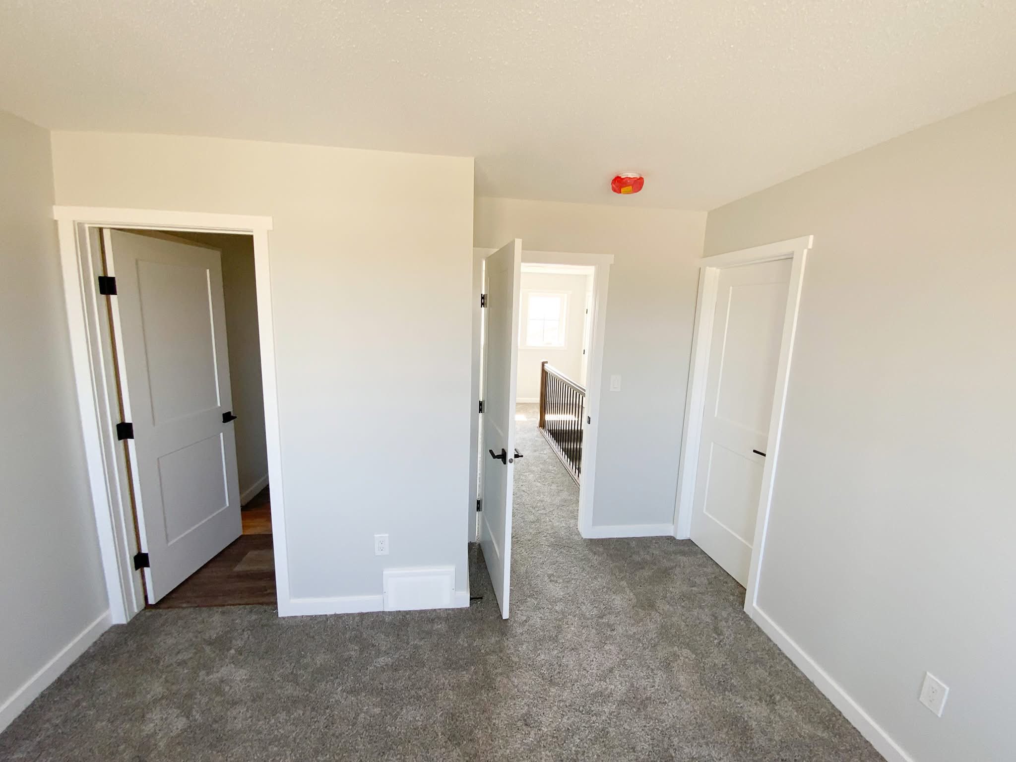Empty room with beige walls and gray carpet, featuring three white doors. One door is slightly open, revealing a hallway with natural light. A red smoke detector is visible on the ceiling. The scene feels neutral and modern.