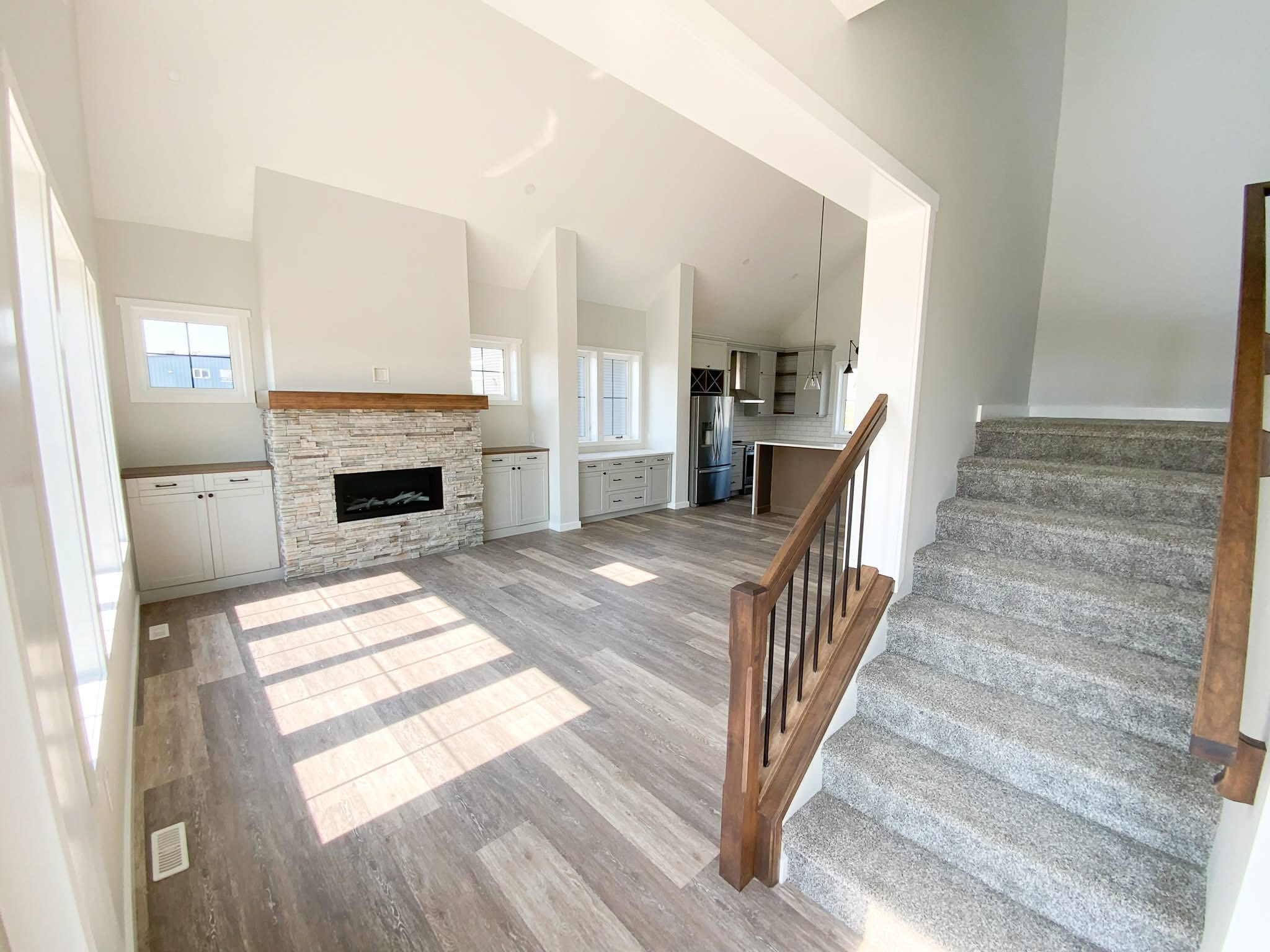 Bright living space with wood flooring and carpeted stairs. Features a stone fireplace, large windows, and adjoining white kitchen. Airy and modern.