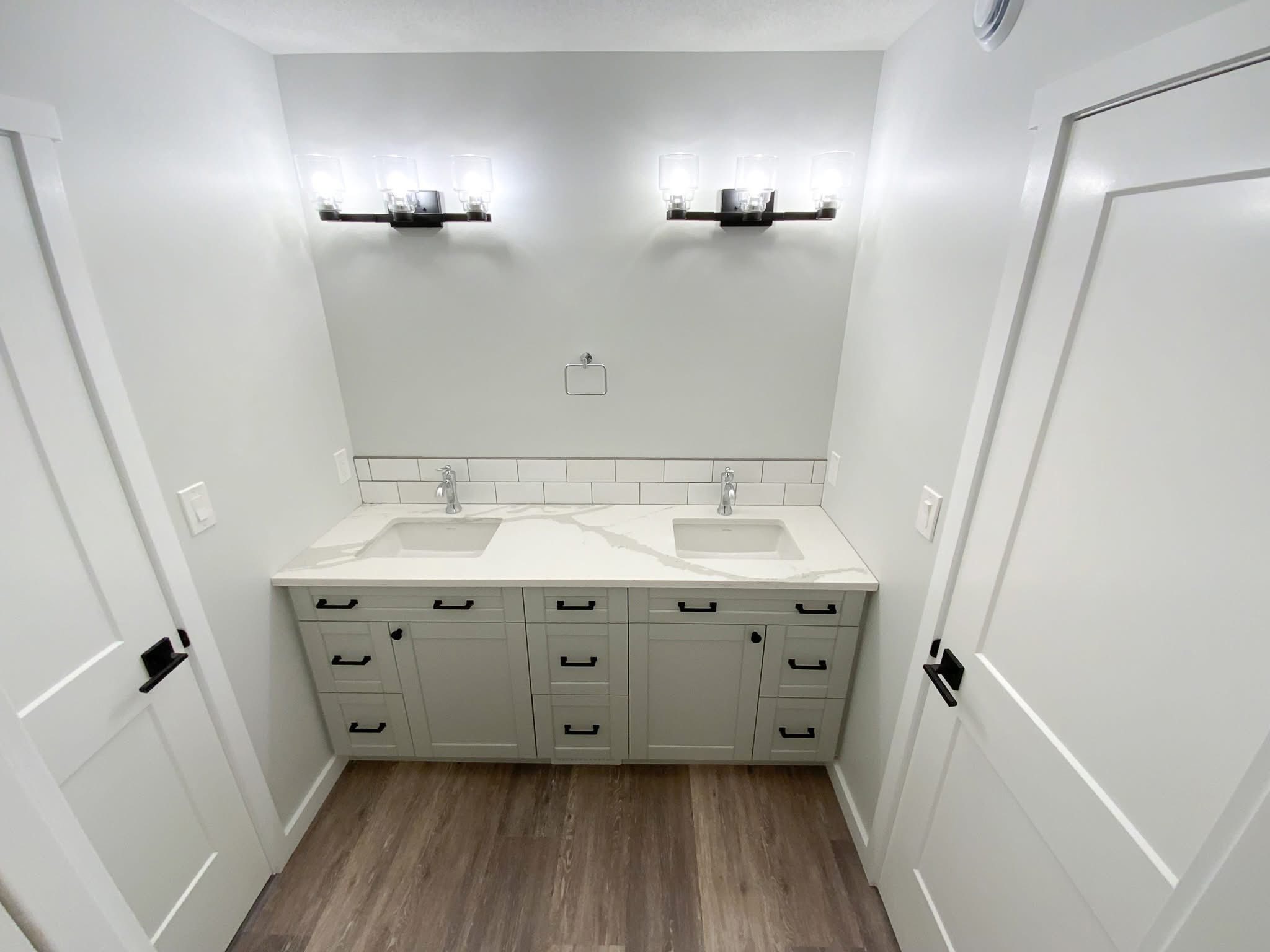 Modern bathroom features a double vanity with light gray cabinets, sleek faucets, and a white countertop. Two bright wall lights illuminate the space, complemented by light wood flooring and white walls, creating a clean and minimalist vibe.