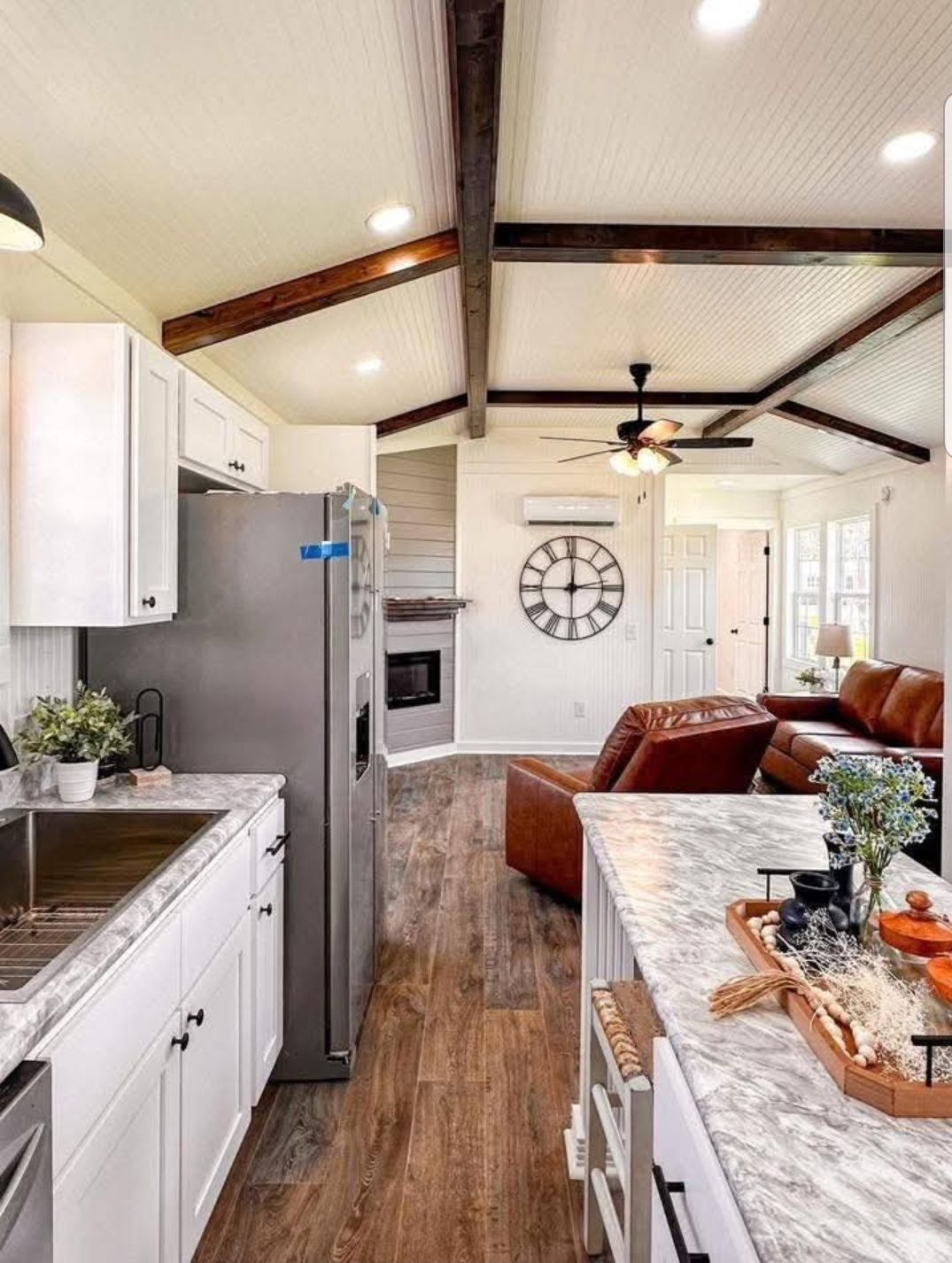 Bright, cozy living space with white cabinets, marble countertops, and wooden floors. Ceiling beams add charm. A clock decorates a wall near a comfy couch.