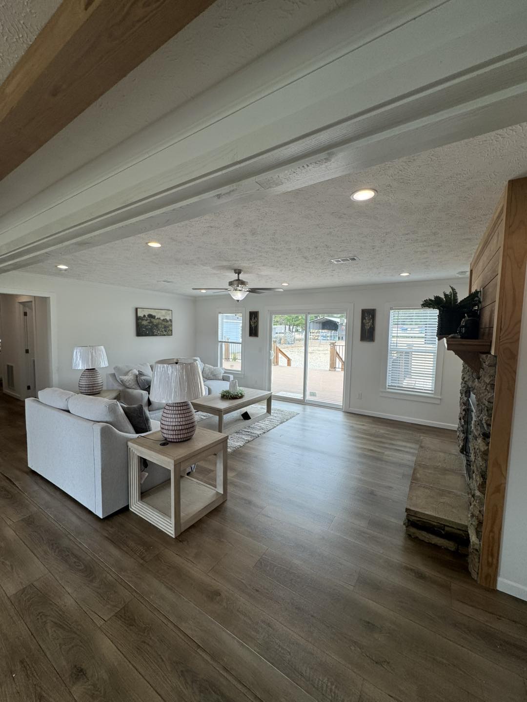 Spacious living room with a cozy ambiance. Features light gray sofas, wooden side tables with lamps, a stone fireplace, and large windows leading to a deck outside.