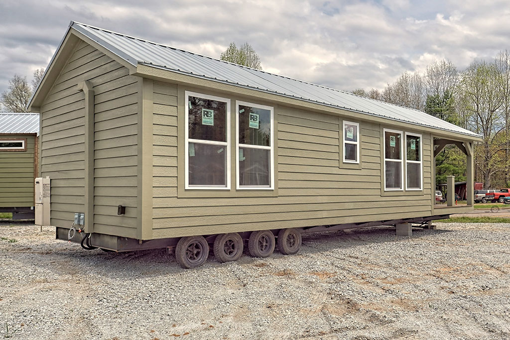 A mobile tiny house with a gray exterior and metal roof sits on a gravel lot. It has multiple windows and is mounted on wheels, with trees in the background.