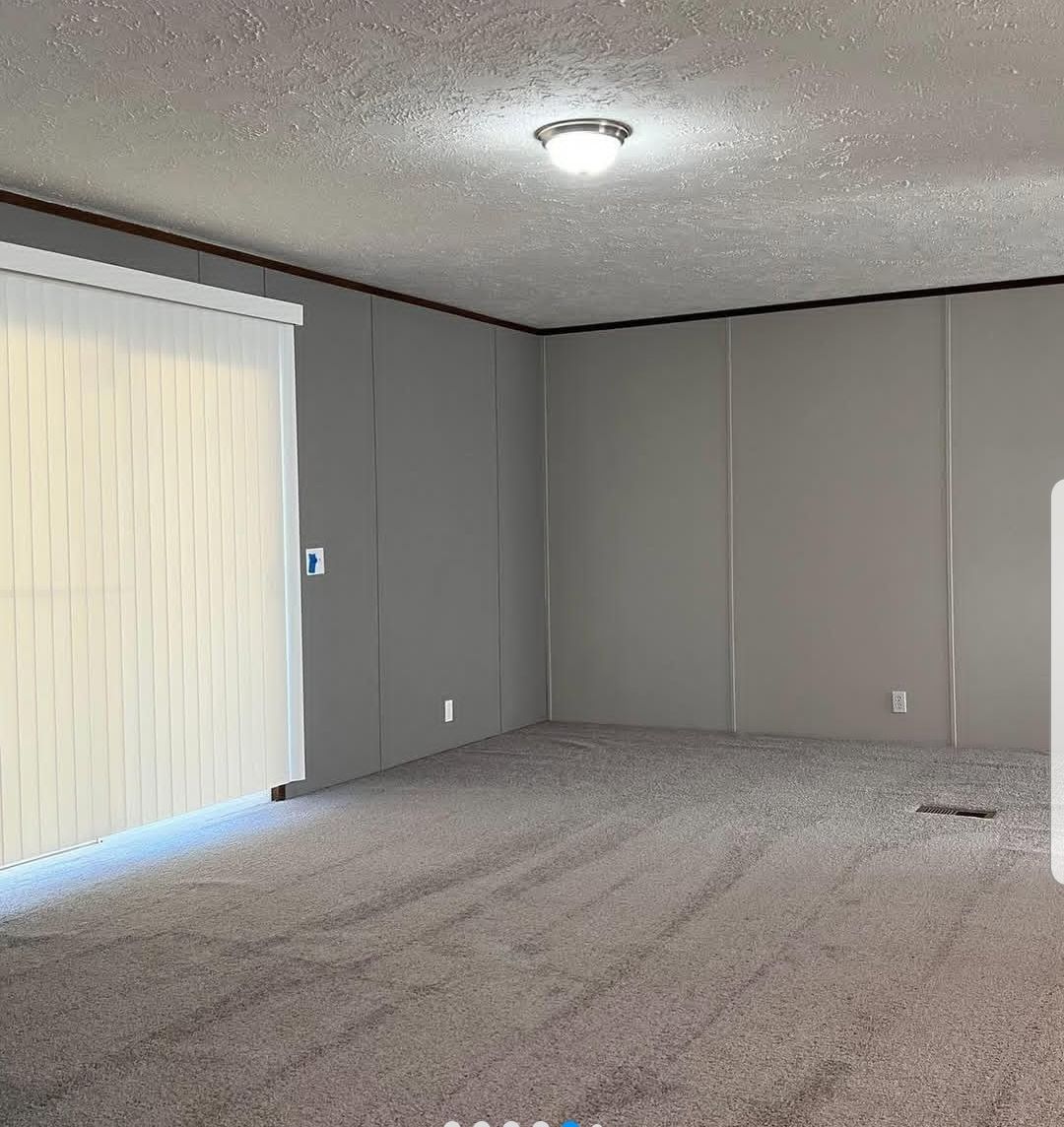 Empty room with gray carpet and walls, illuminated by a ceiling light. A large window with vertical blinds is partially open, creating a calm ambiance.
