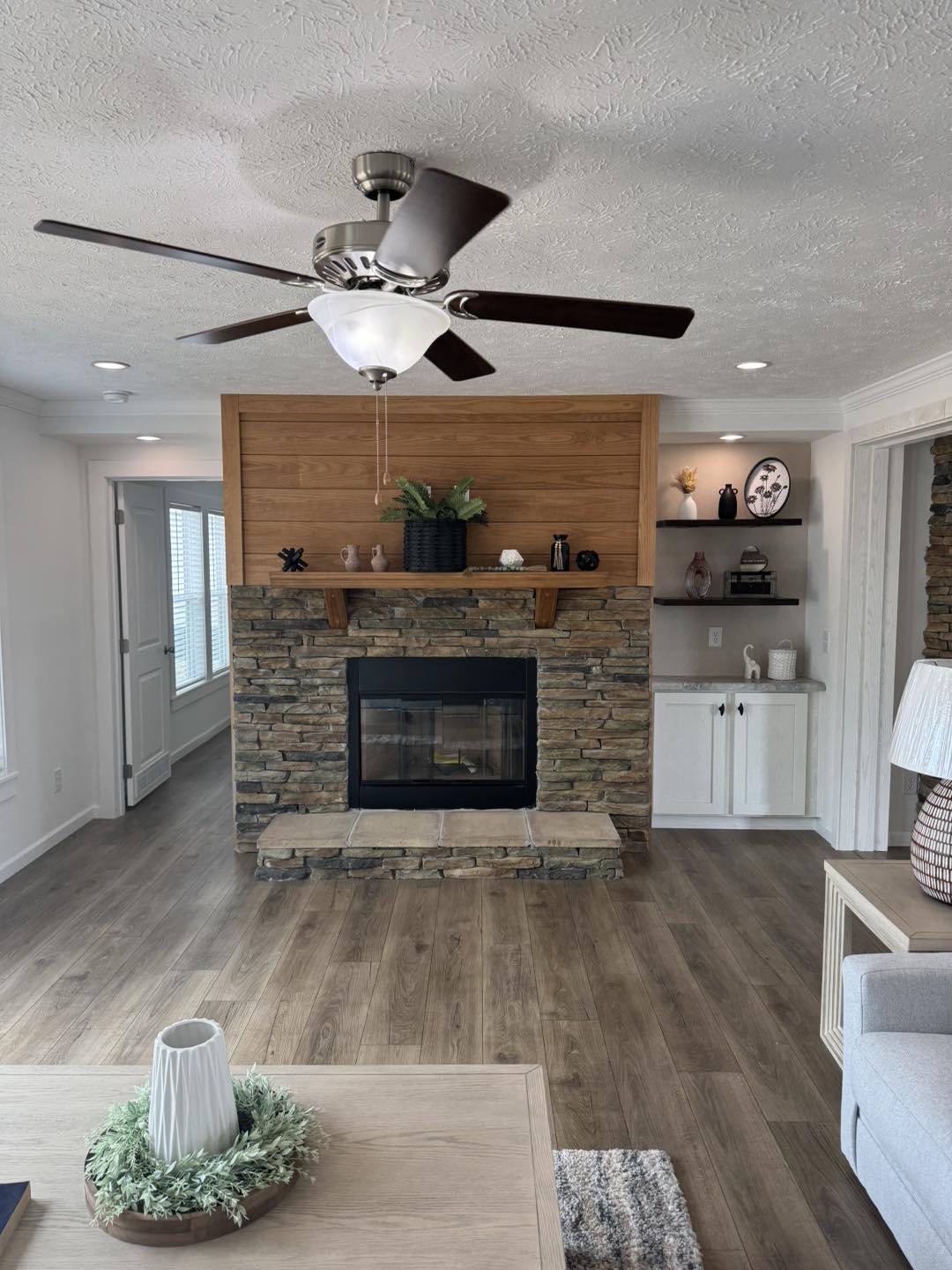 Cozy living room with a stone fireplace and wooden mantel, adorned with plants and decor. A ceiling fan hangs above, flanked by shelves and soft lighting.
