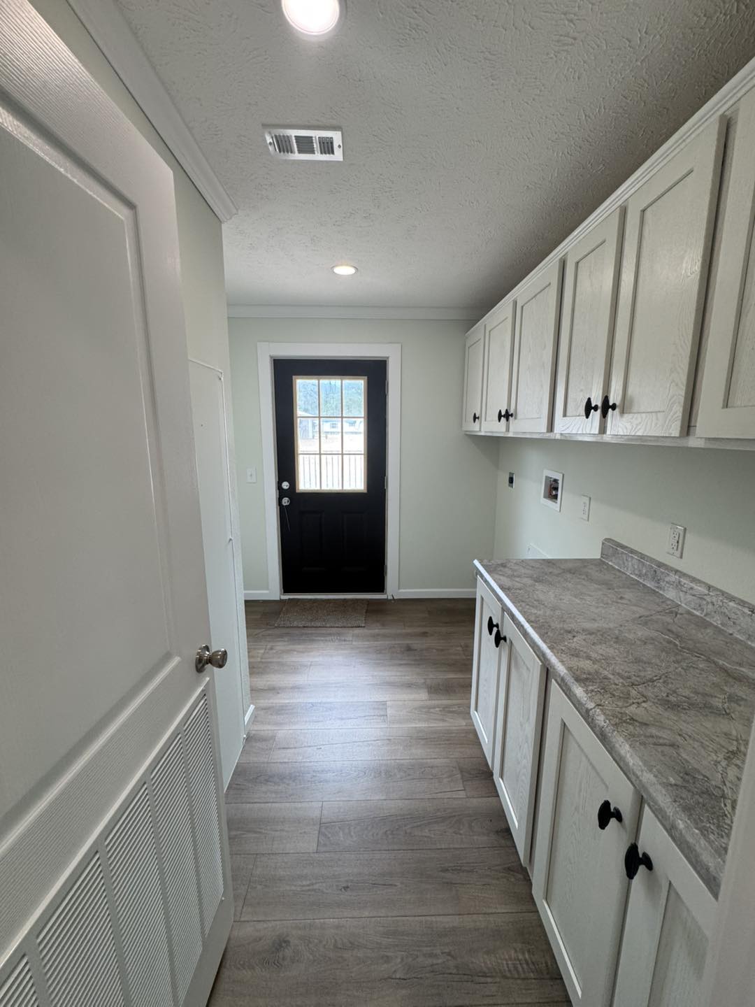A bright laundry room with white cabinets and a marble countertop on the right. Light wood flooring leads to a black door with a window, creating an inviting tone.