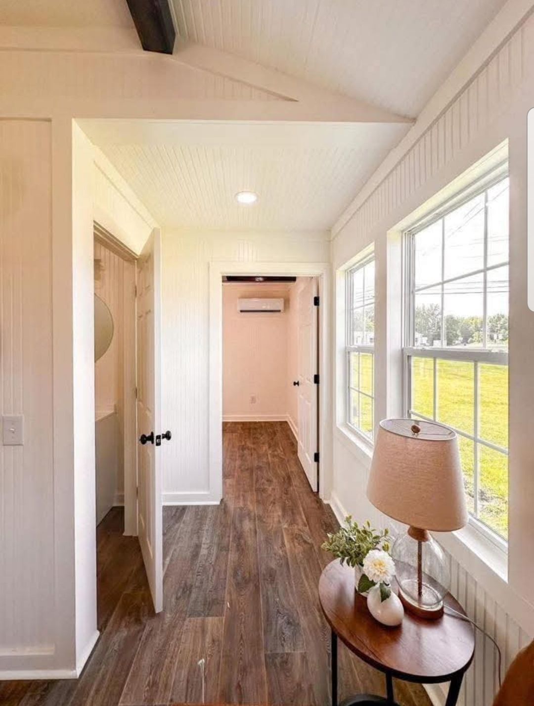 Narrow hallway with wood floors, white walls, and large windows on the right. A round table holds a lamp and flowers, creating a warm, inviting tone.
