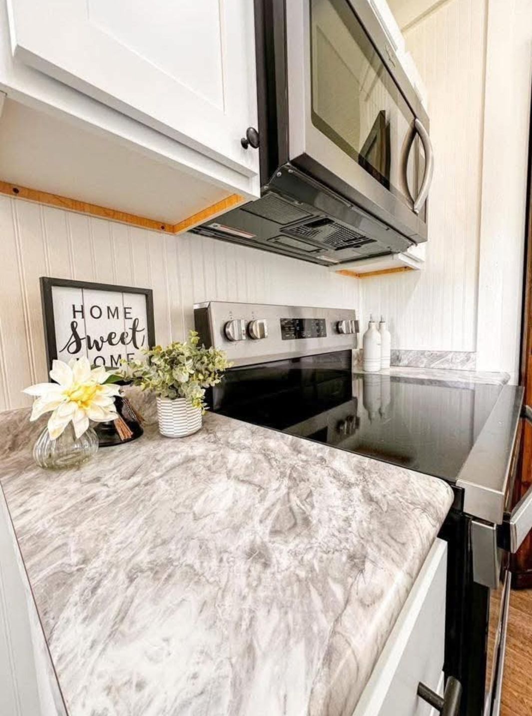 Compact, modern kitchen corner with marble countertop, silver stove, and microwave. Decor includes small plants, white flowers, and a "Home Sweet Home" sign, creating a cozy ambiance.
