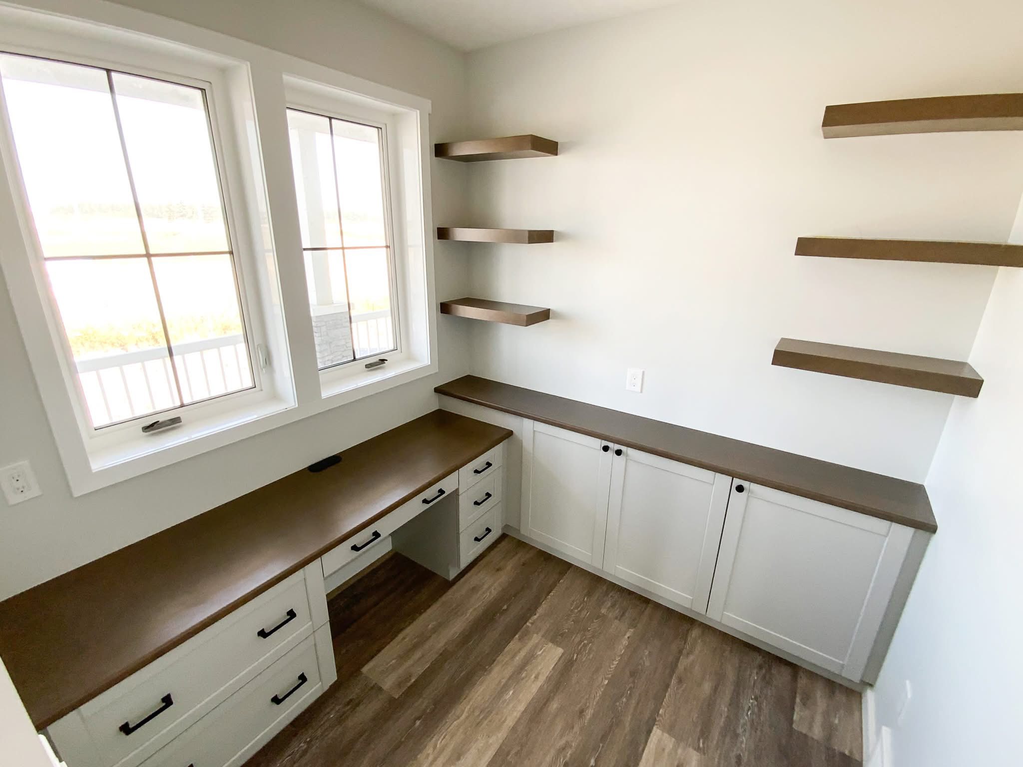 Modern home office with dark wood countertops, white cabinets, open shelves, and two windows. Light wooden floor adds a minimalist touch.