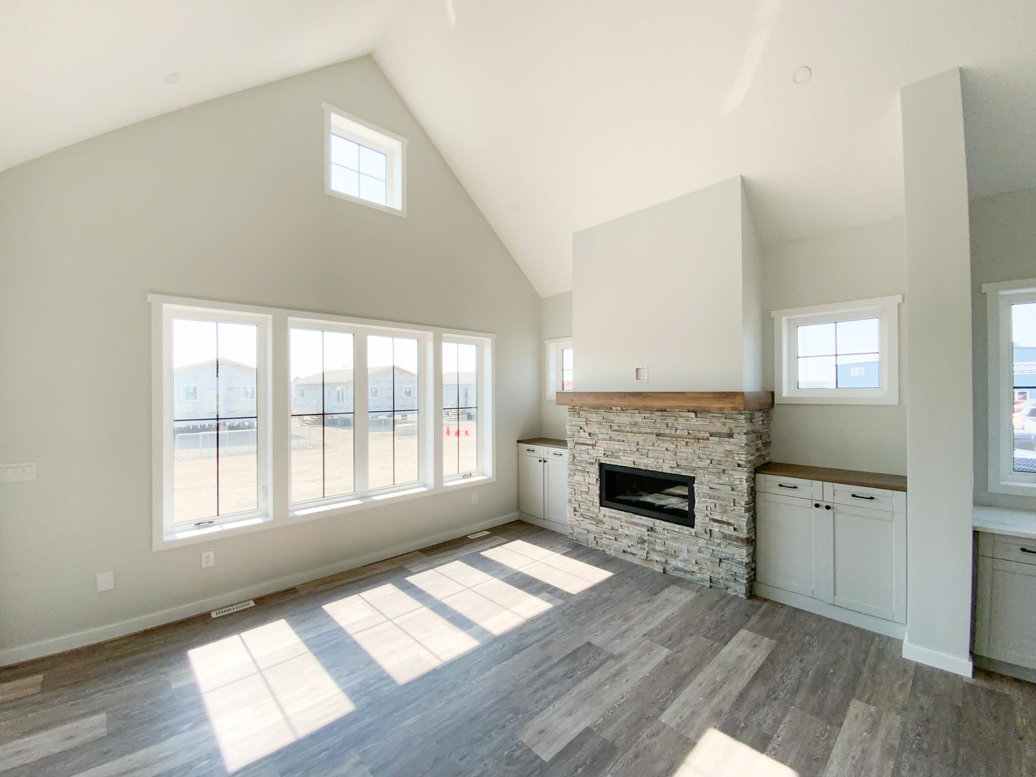 Spacious, bright living room with high ceilings, large windows, and sunlight streaming in. Stone fireplace on right, flanked by built-in cabinets. Modern and airy atmosphere.