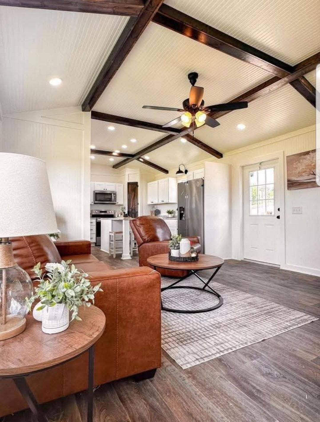 A cozy living room with a brown leather couch and armchair. Wooden beams, a ceiling fan, and rustic decor create a warm, inviting atmosphere.