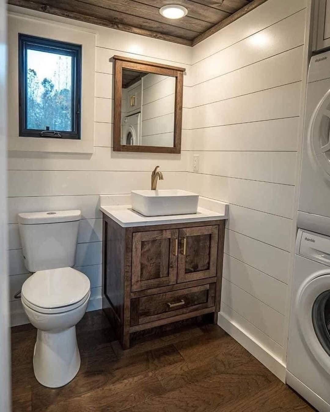 Cozy bathroom with wood panel walls, a rustic wood vanity with a vessel sink, next to a toilet. A washer-dryer is tucked beside the vanity. Natural light filters in.