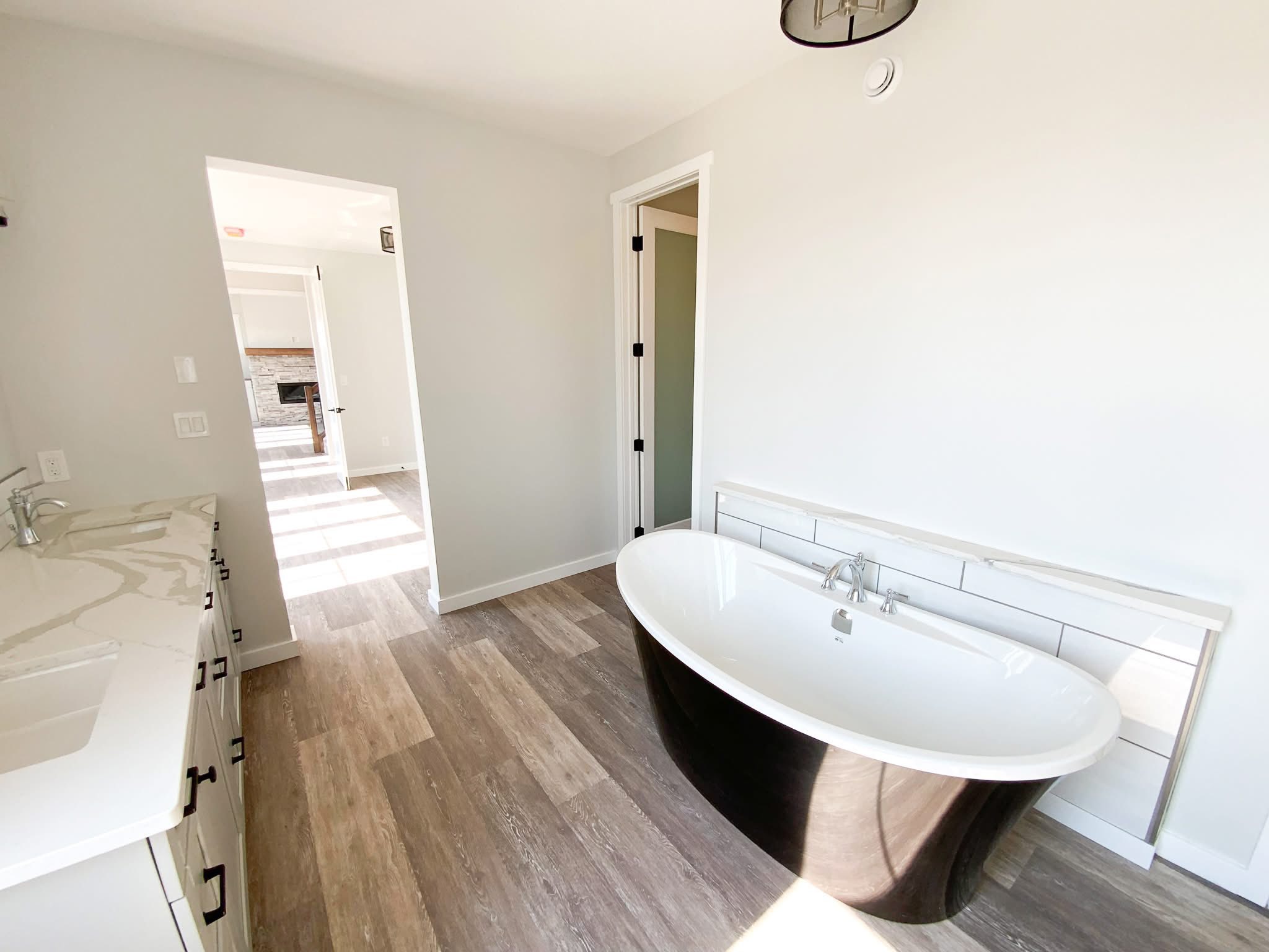 Bright bathroom with a modern stand-alone black bathtub on wooden flooring. White countertops with dual sinks are on the left, creating an airy atmosphere.