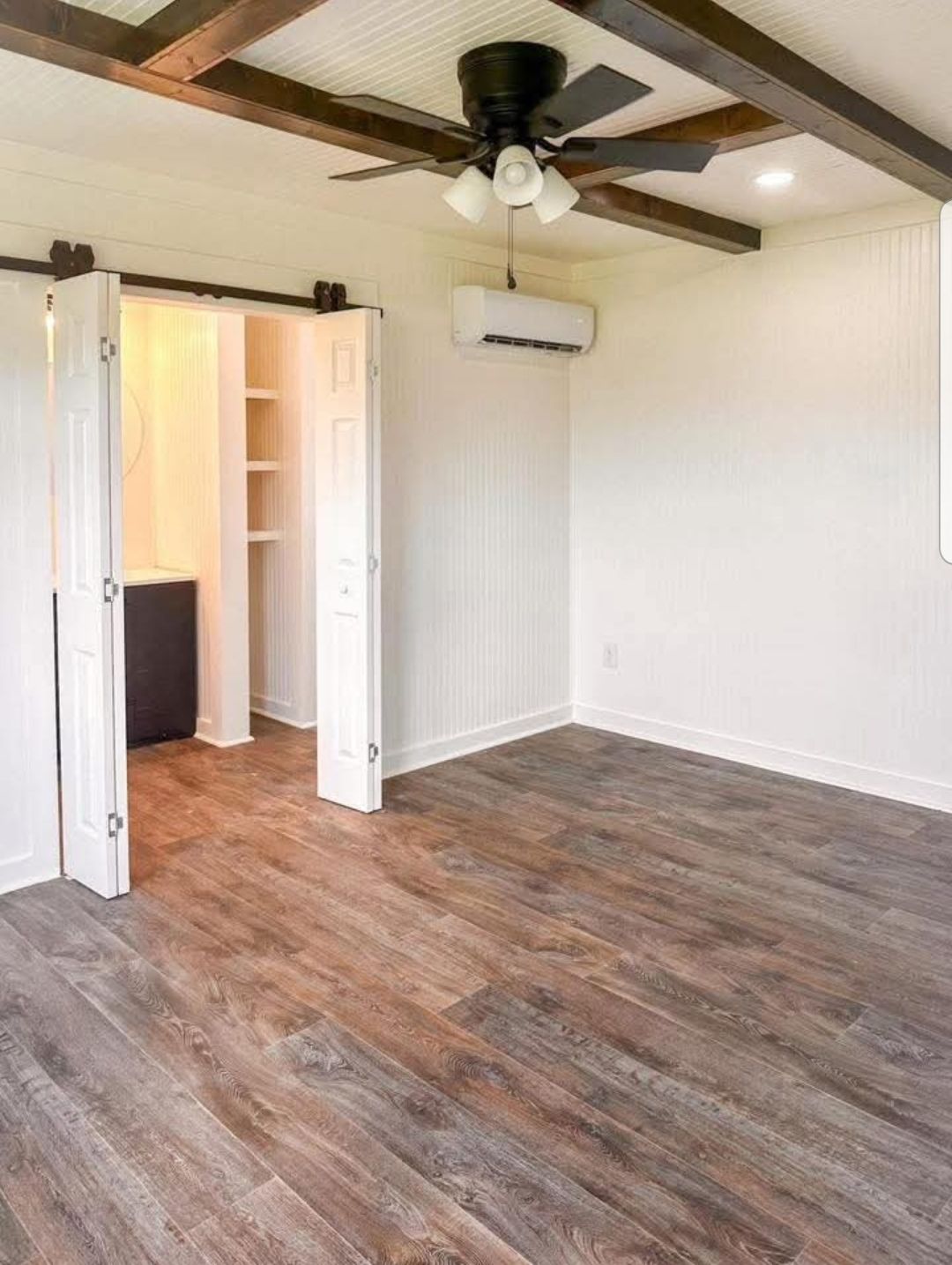 A bright room with wooden floors, white walls, a ceiling fan with exposed beams, and an open door revealing a storage area. Airy and minimalistic.