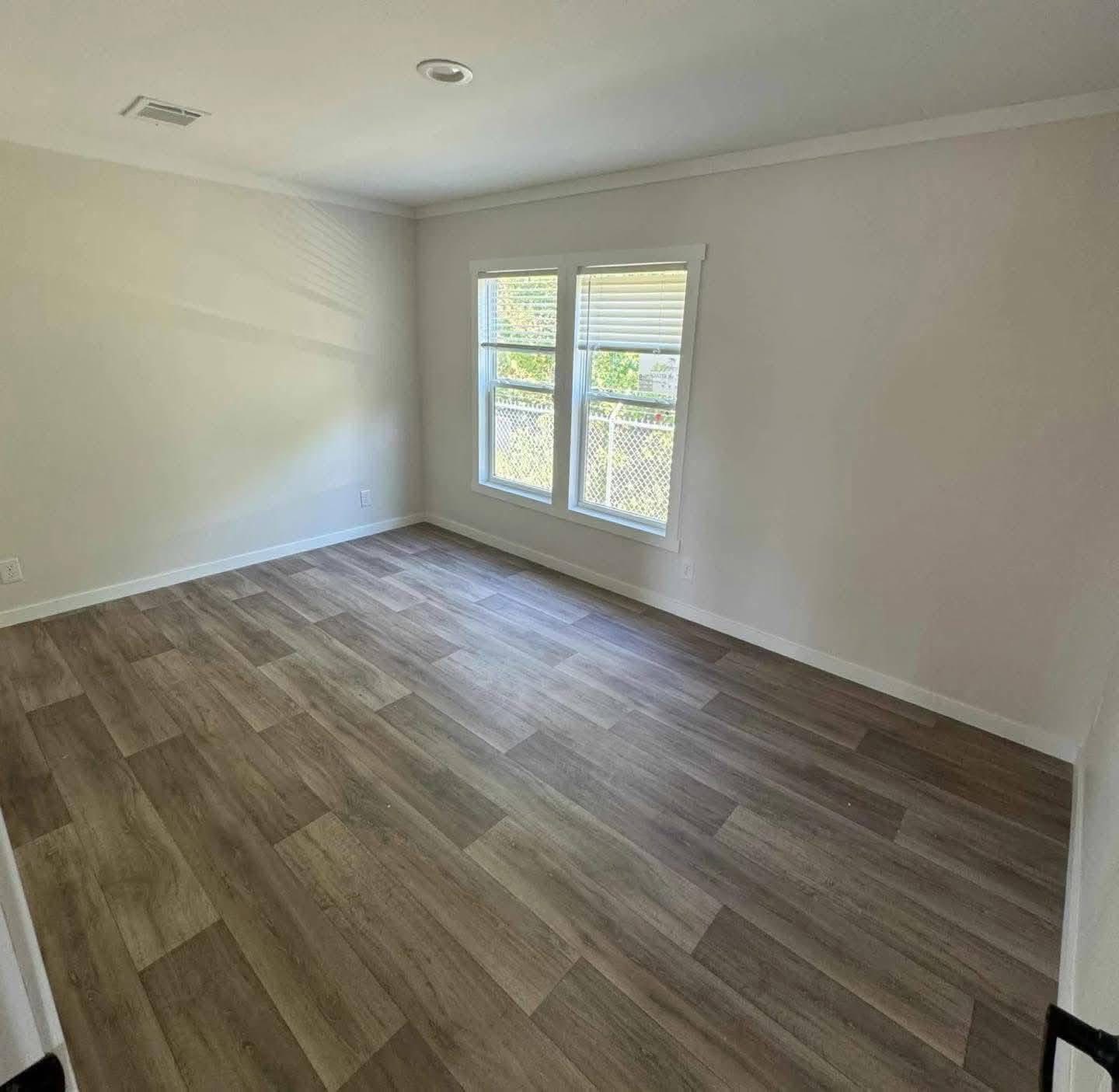 Empty room with light wood vinyl flooring, beige walls, and a double window allowing natural light. The atmosphere is bright and minimalistic.