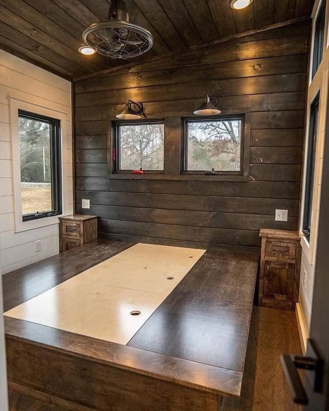 Cozy, rustic room with dark wooden walls and ceiling, featuring a wooden platform bed. Two small windows bring in soft natural light, enhancing the warm ambiance.