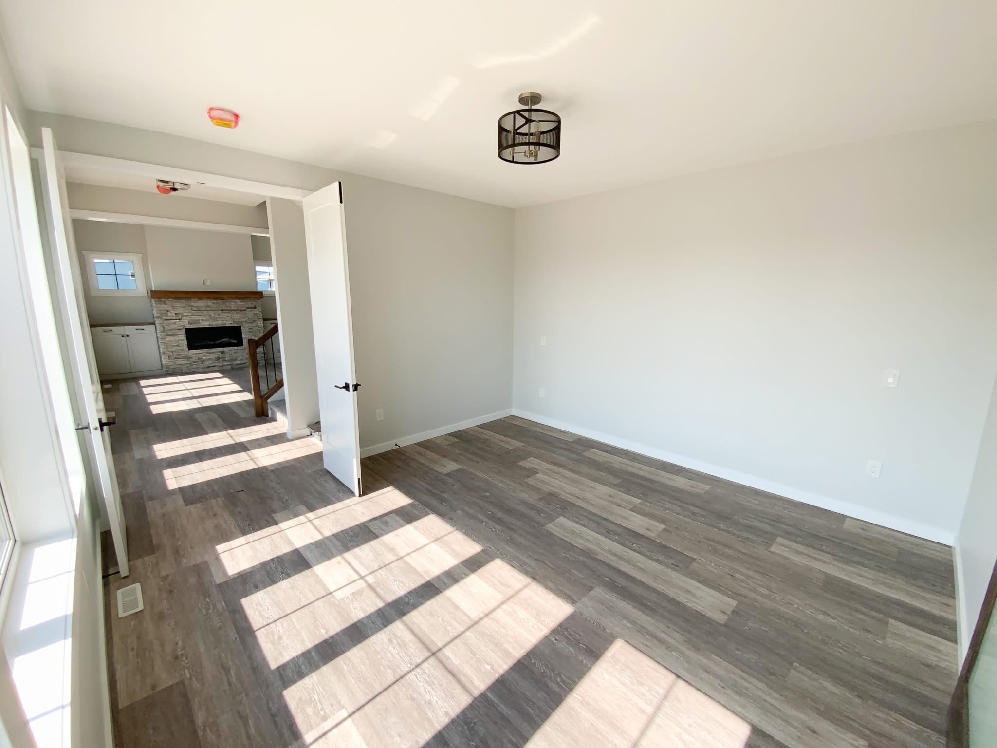 Bright, empty room with light gray walls, wooden floor, and sunlight streaming through a large window. An open door leads to a room with a modern fireplace.