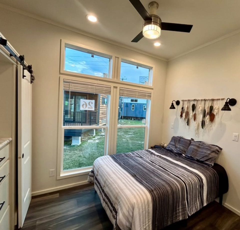 Cozy bedroom with a striped bedspread, large windows, and a ceiling fan. Feather decor hangs above the bed, creating a calm, inviting atmosphere.