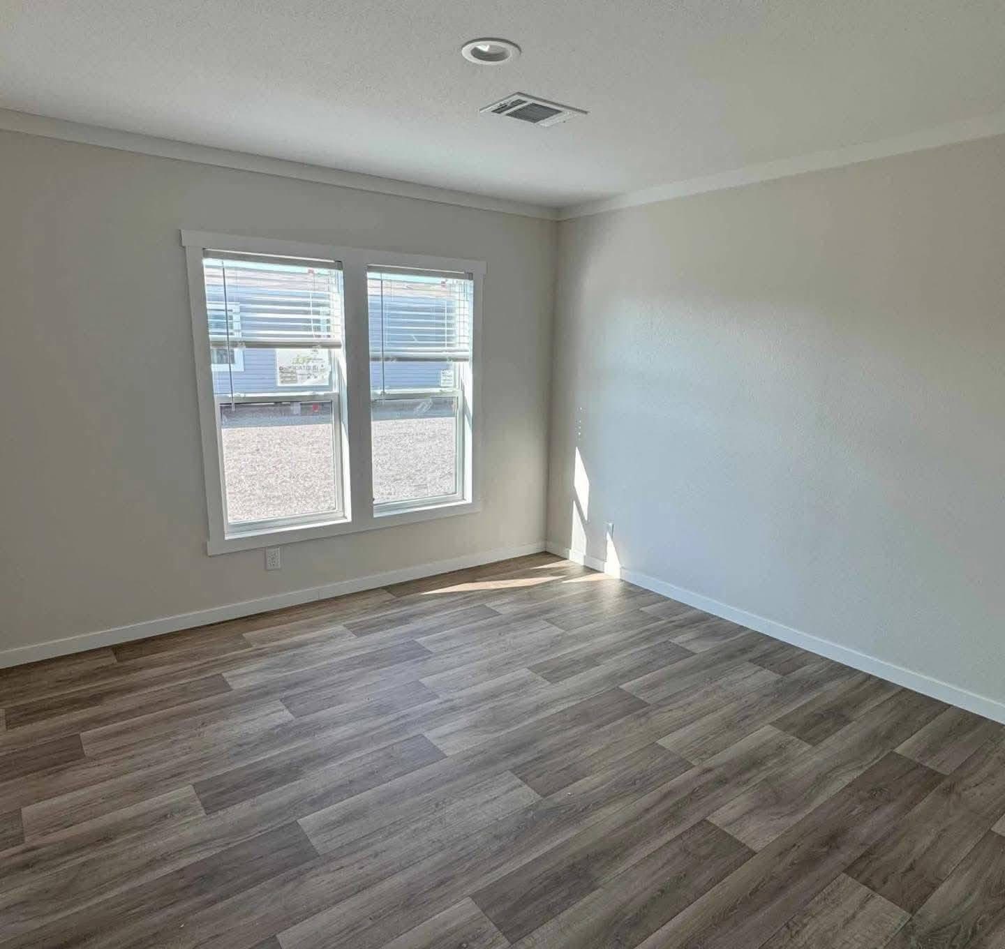 An empty room with light gray walls and wood-patterned flooring. Sunlight streams through a double window, creating a calm, inviting atmosphere.