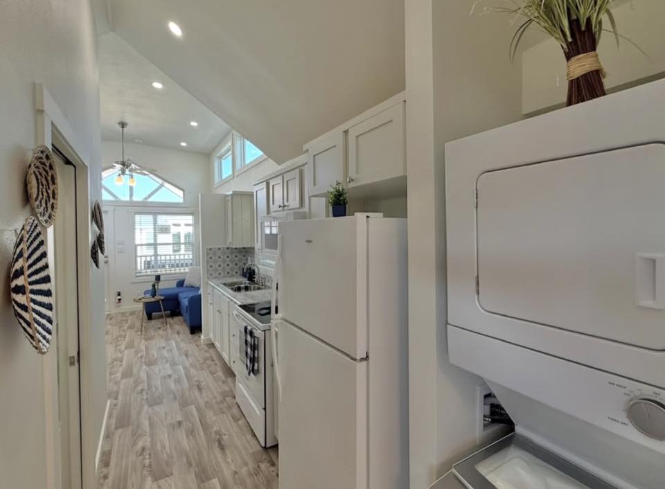 Bright, narrow kitchen with white cabinets and appliances. Laundry unit on the right, living area with blue sofa and large window at the end.