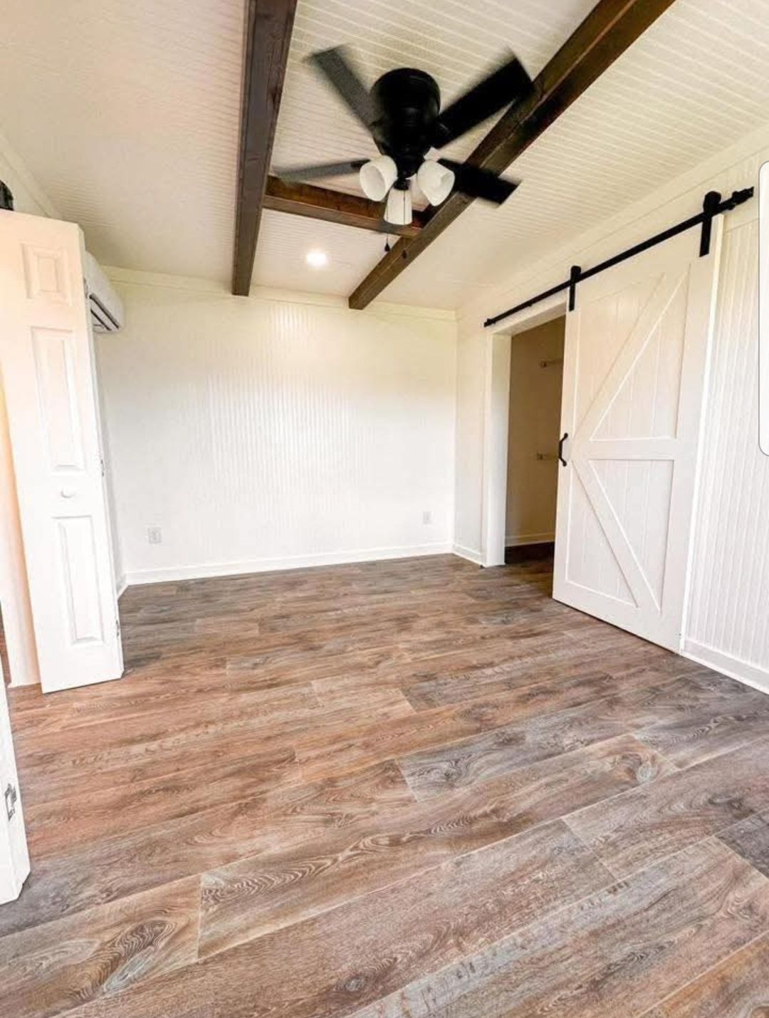 An empty room with wooden flooring, white walls, and exposed wooden beams on the ceiling. A black ceiling fan is centered, and a white barn door is on the right.