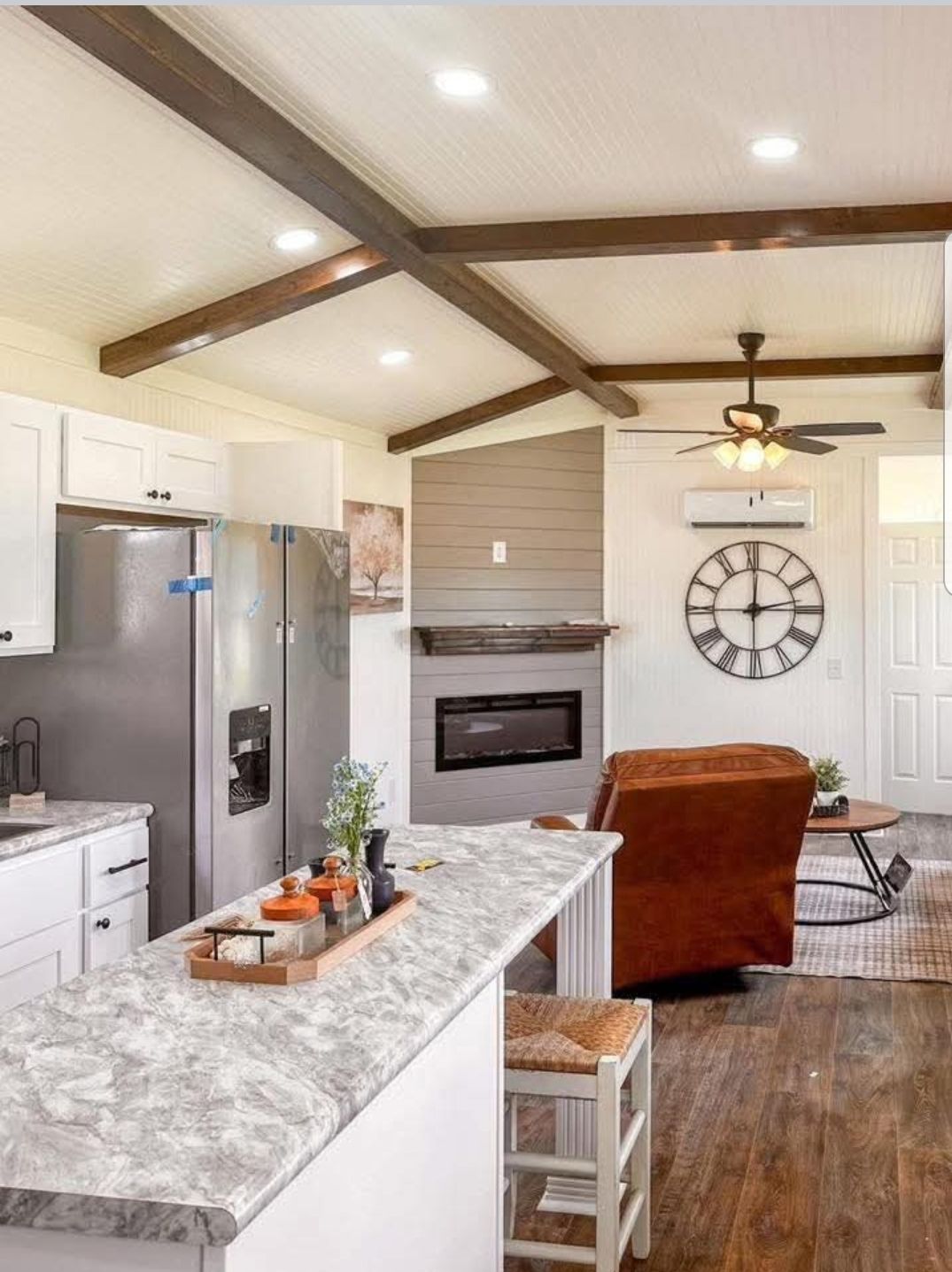 Modern kitchen and living area with wood beams and stone countertops. Brown leather chair faces a fireplace. Large wall clock adds rustic charm.