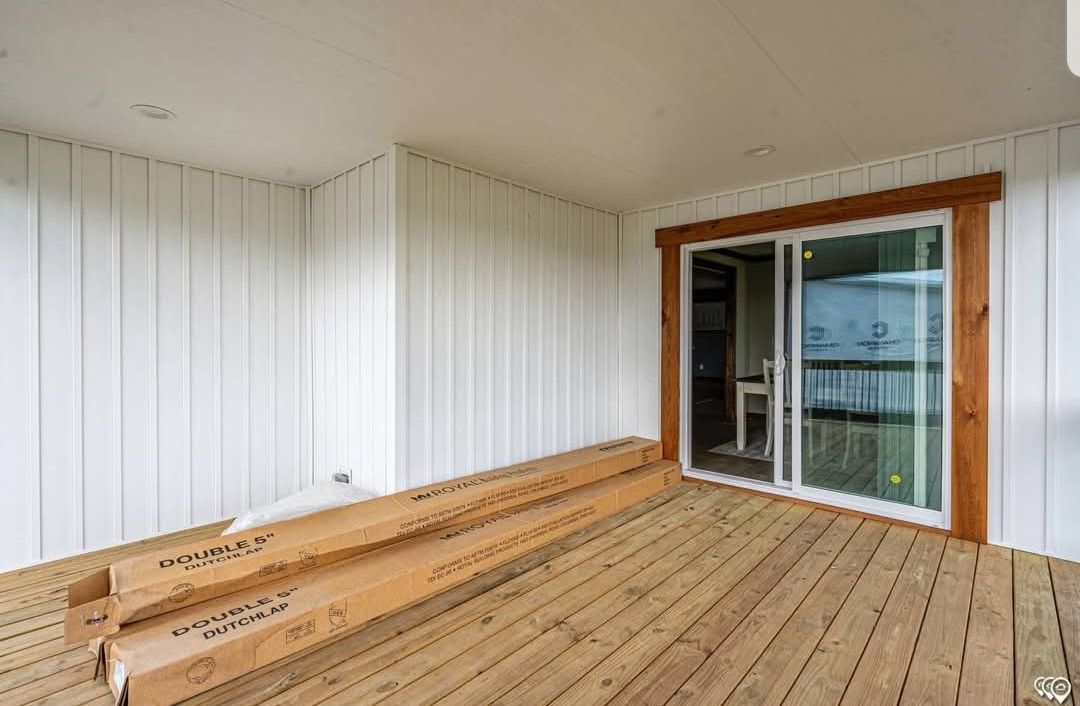 A wooden deck with unfinished timber planks, white paneled walls, and a glass sliding door framed in wood. Cardboard boxes labeled "Double 5" Dutchlap" are on the floor.