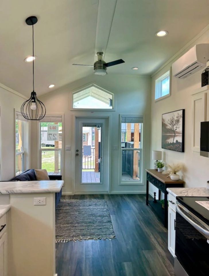 Compact, modern living room with high ceiling, fan, and pendant light. Wooden floor, neutral tones, art on wall. Glass doors lead to patio, creating airy feel.