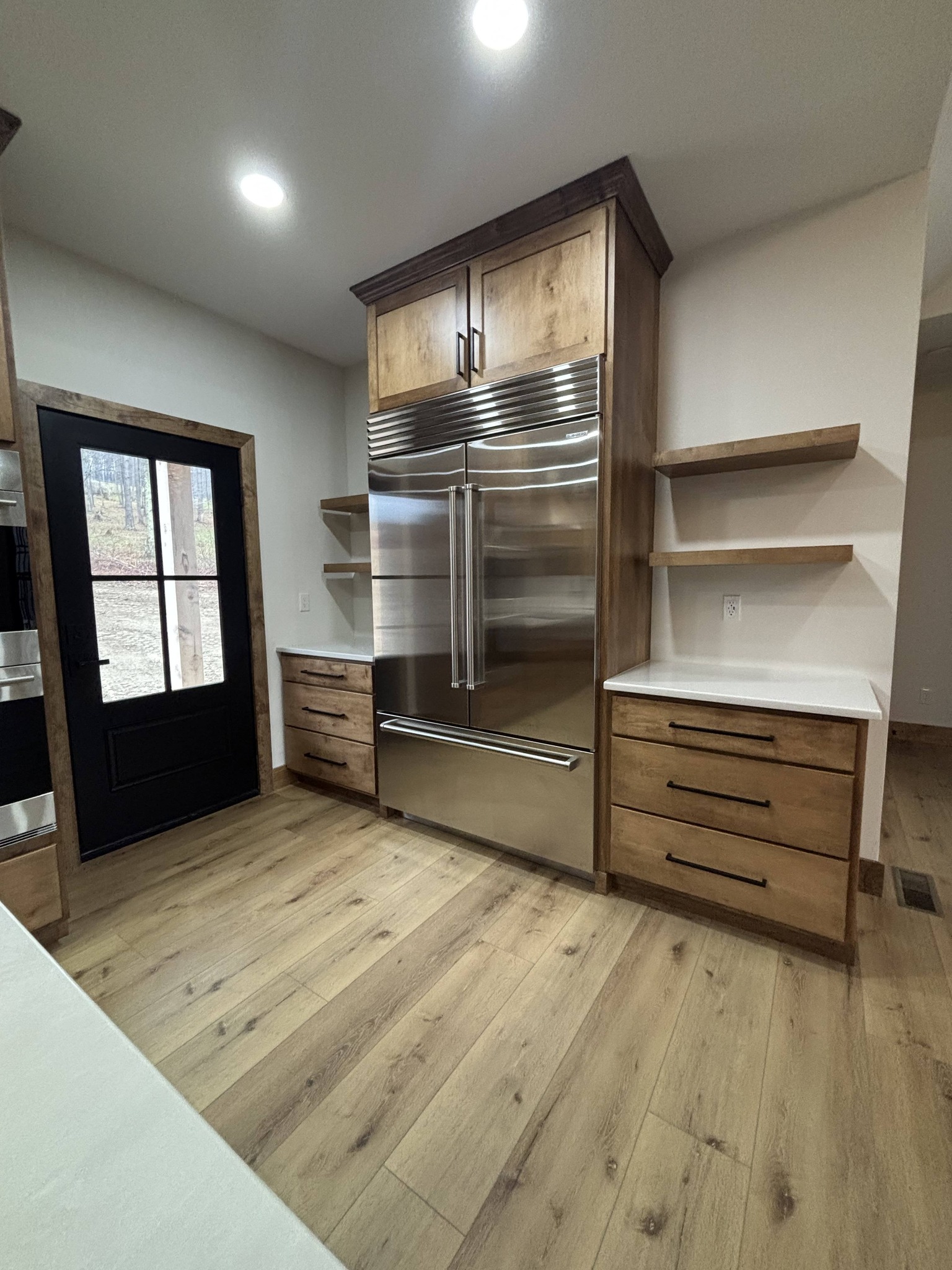 Modern kitchen with wood flooring, a large stainless steel refrigerator, wooden cabinets, white countertops, floating shelves, and a glass-paneled black door. Bright lighting creates a clean, welcoming atmosphere.