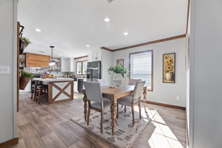 A bright, modern kitchen and dining area with wooden floors and light gray walls. Sunlight streams through a window, illuminating a wooden dining table with four upholstered chairs. A vase with greenery sits on the table. The kitchen features a large island with stools, pendant lights, and stainless steel appliances. A sunflower painting adds a pop of color to the room.
