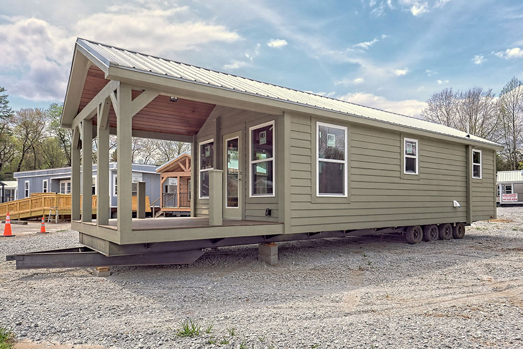 A light green tiny house on wheels with a metal roof and porch sits on gravel under a blue sky. Nearby, another tiny house and orange cones are visible.