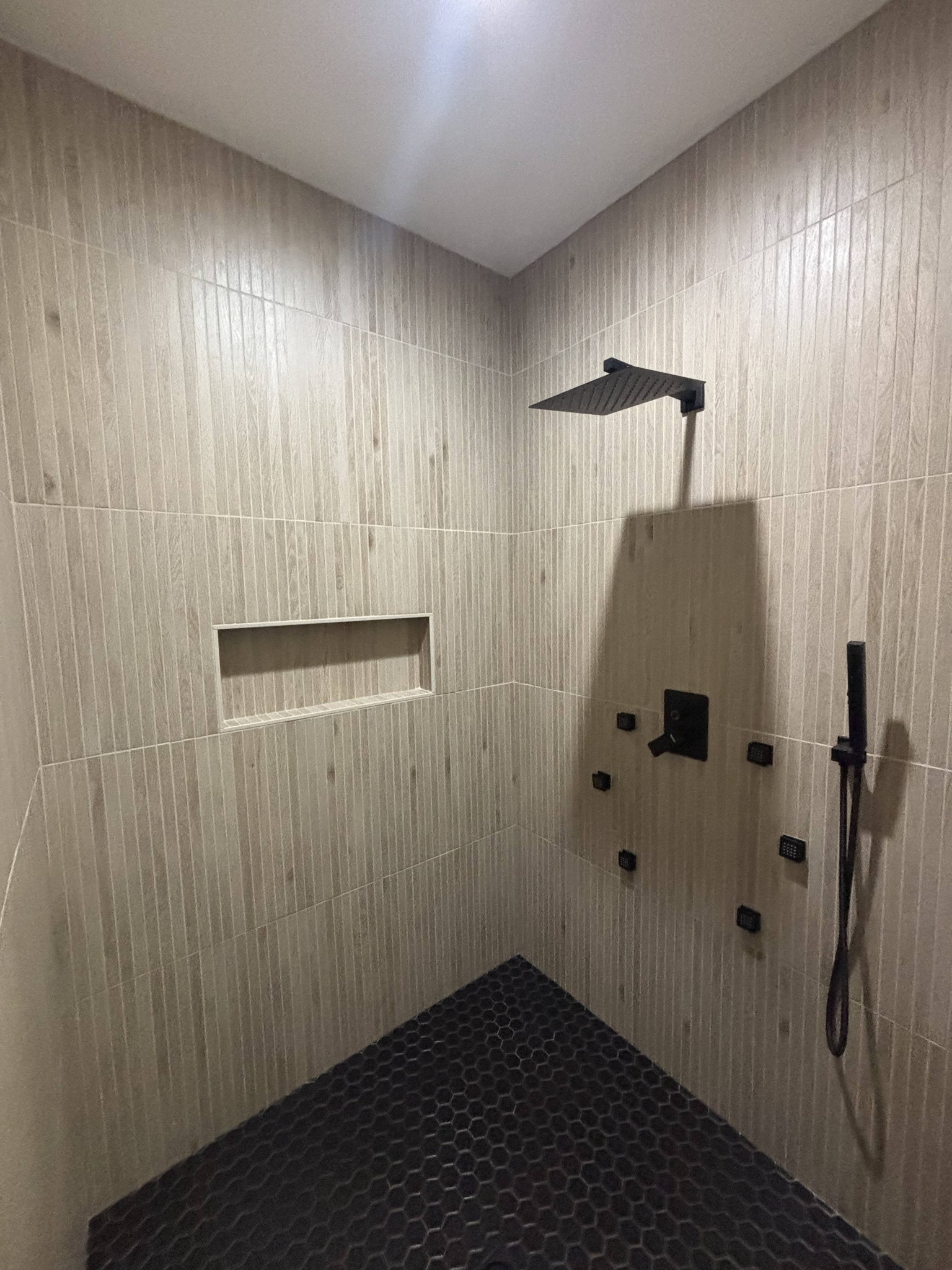 Modern shower with beige vertical tile walls and dark hexagonal floor tiles. Features a square rain showerhead and a built-in wall shelf.